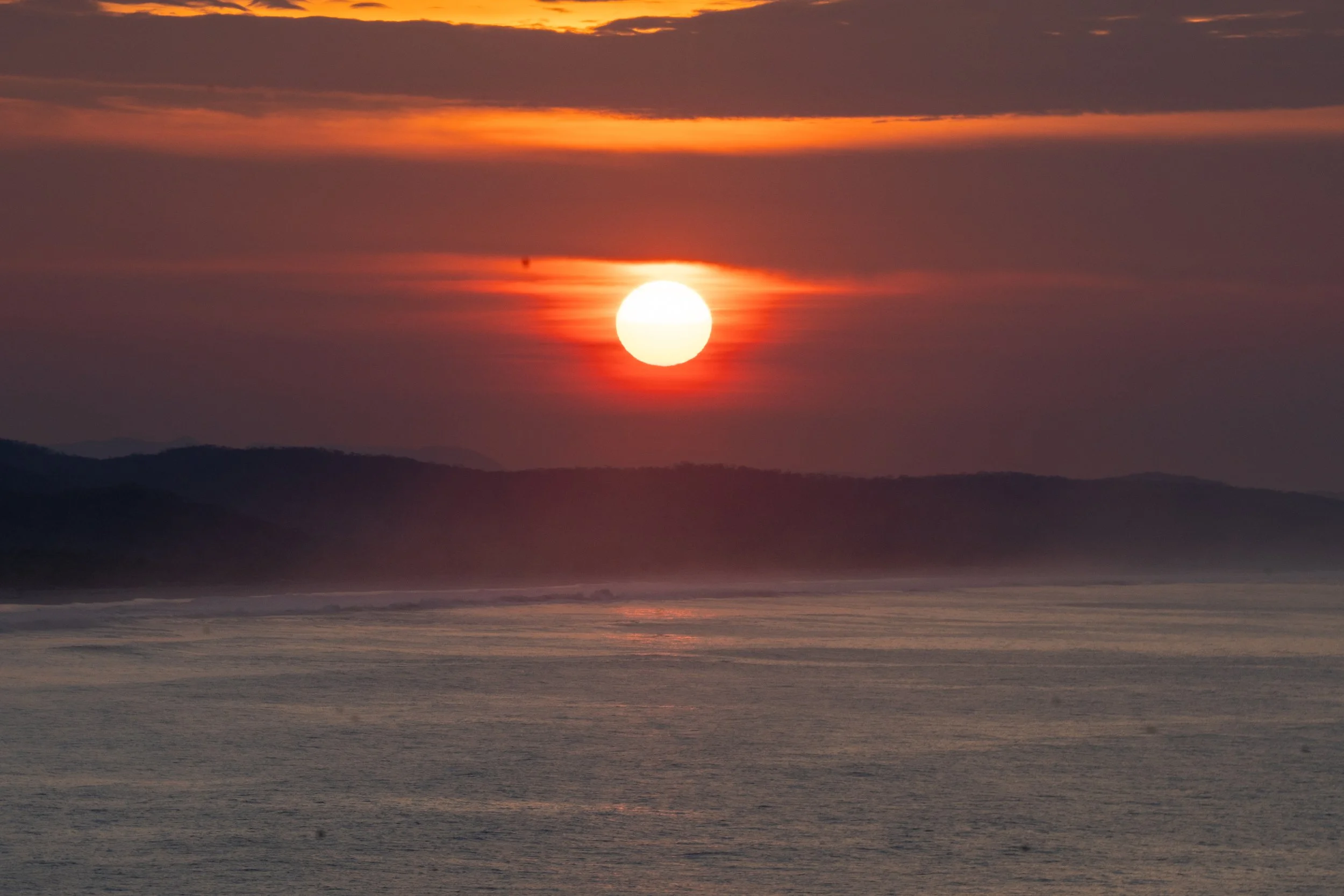 A sunset over a body of water with clouds in the sky, distant hills or mountains, and the sun partially obscured by clouds.