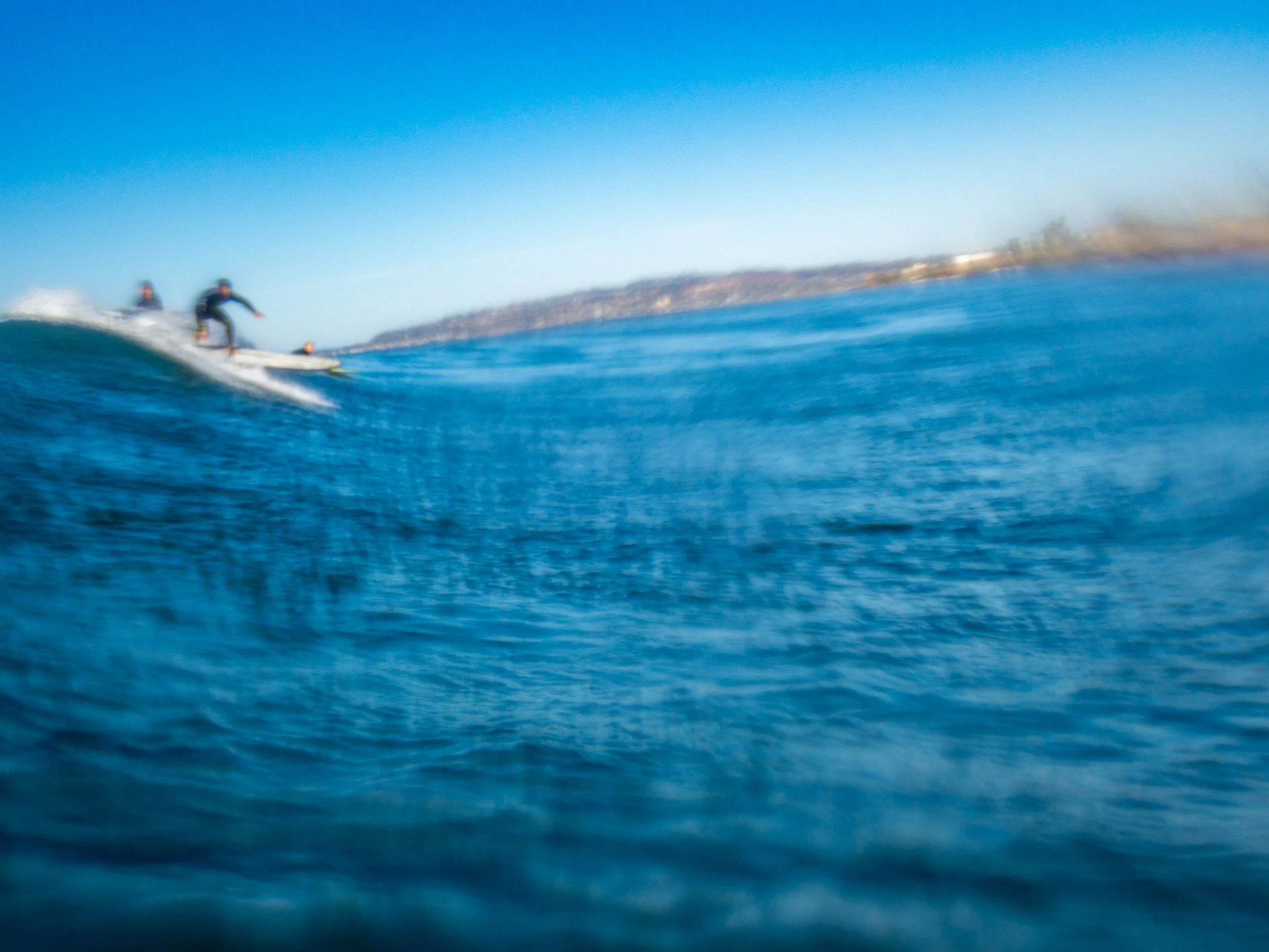 Two surfers riding a wave in the ocean under a clear blue sky with a distant shoreline on the horizon.