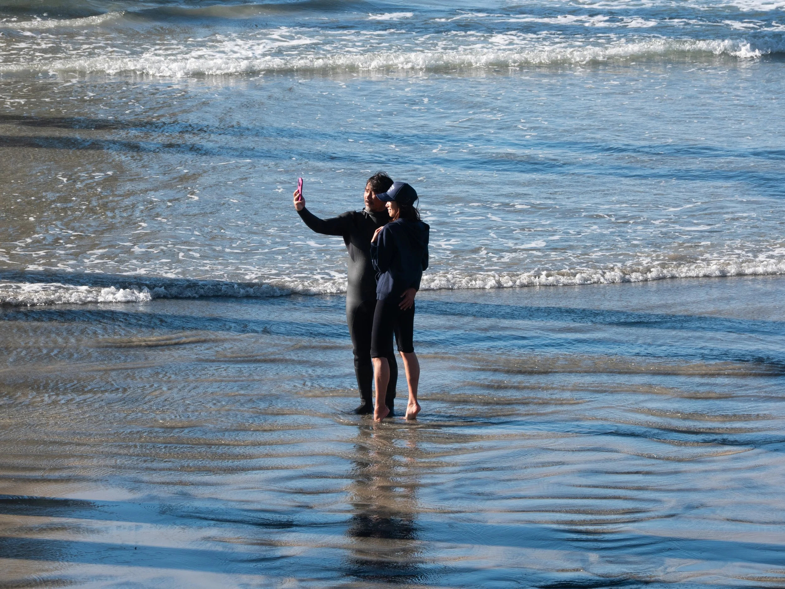 Two people taking a selfie on the beach with ocean waves in the background.