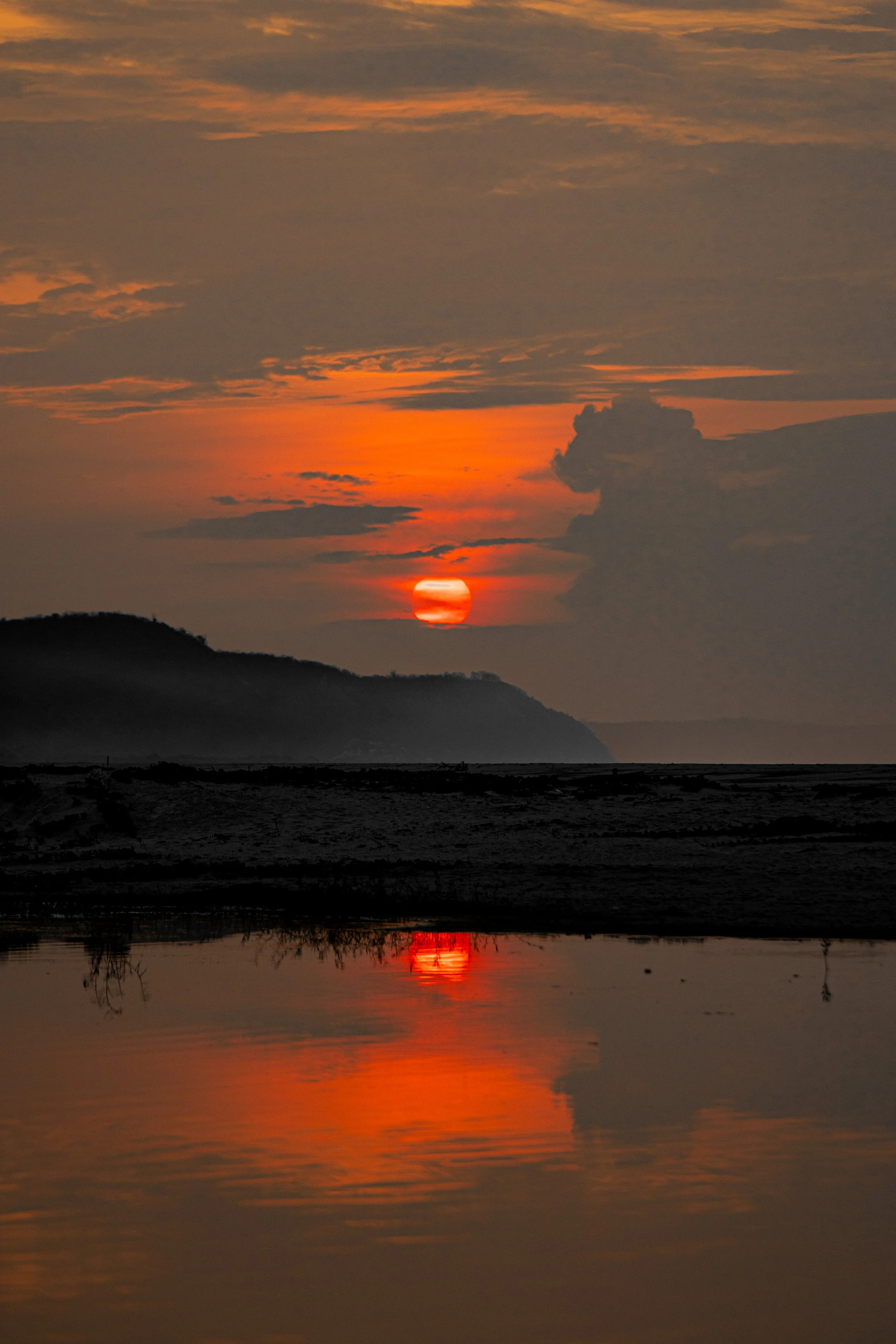 Sun setting over a mountain landscape with clouds in the sky, reflected in calm water below.