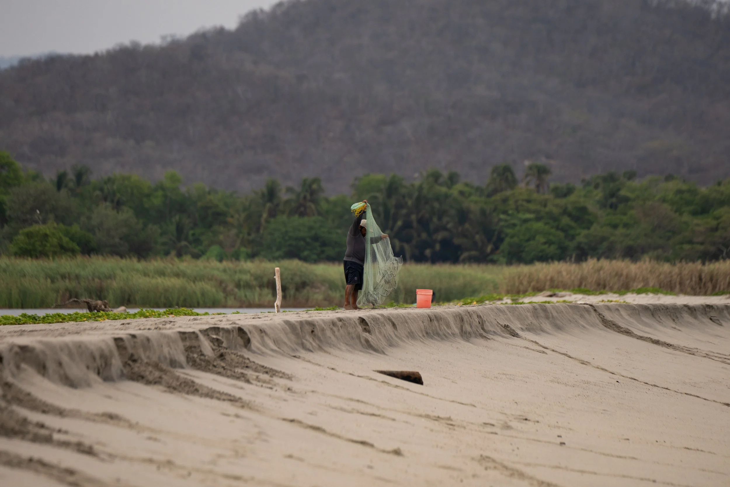 A person standing on the edge of a canal or waterway holding a fishing net. The background shows green trees and a mountain with a mostly bare slope. There are some orange buckets on the ground nearby.