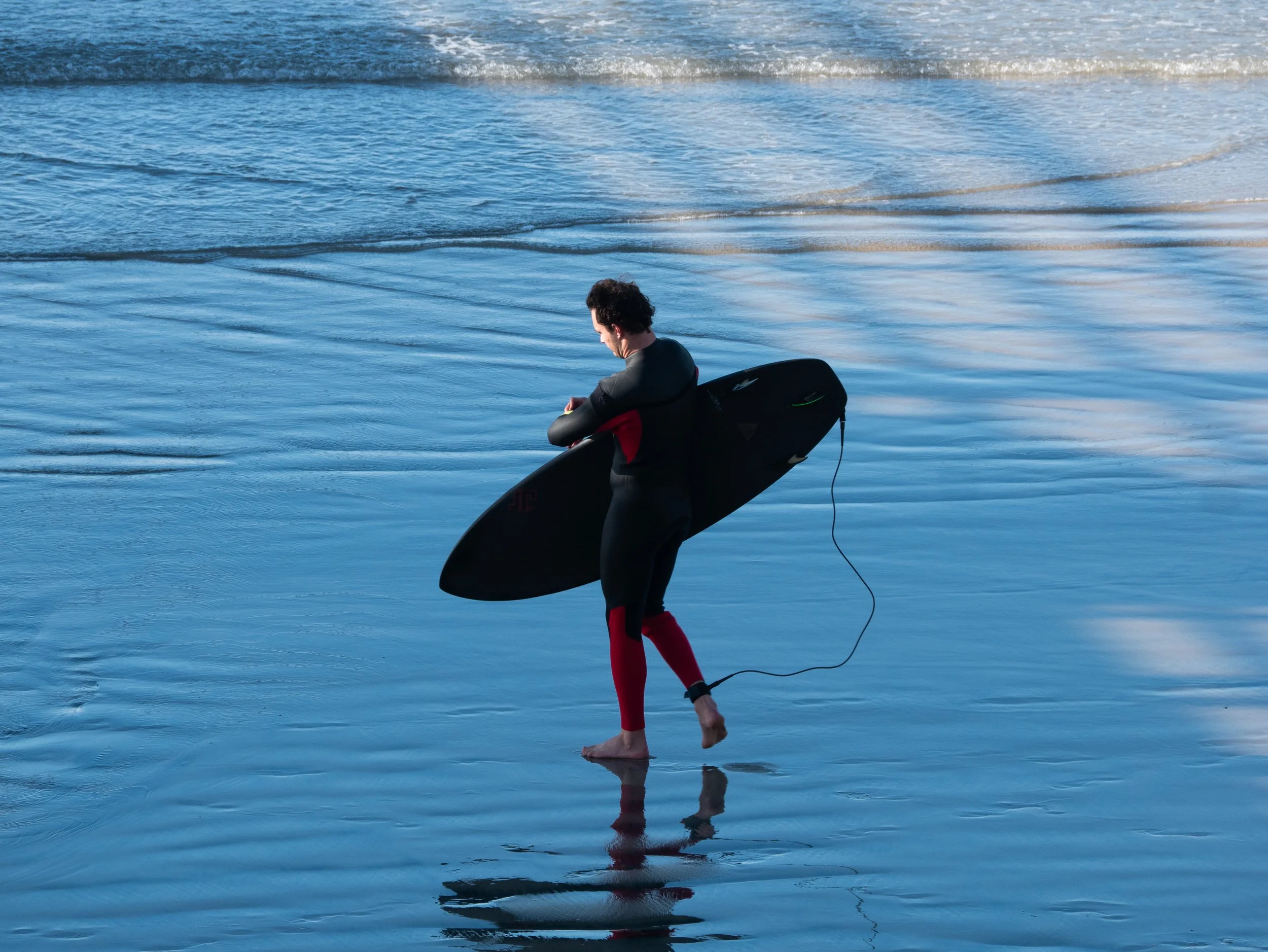 Person in wetsuit carrying a surfboard walking in shallow ocean water.