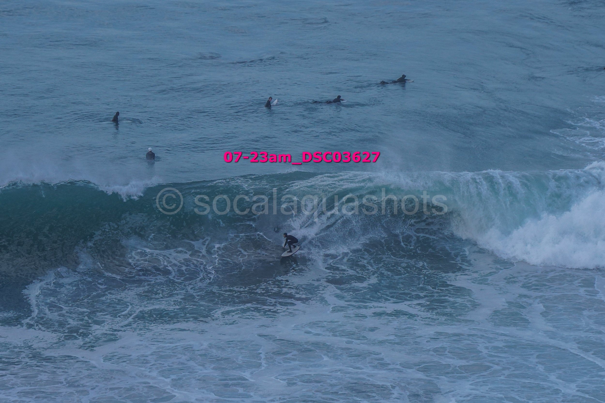 A person surfing on a wave in the ocean with five surfers in the background, some on stand-up paddleboards.