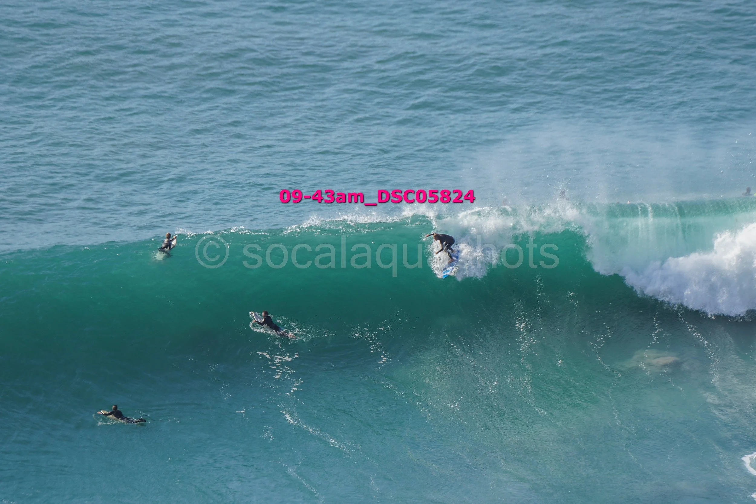 Four surfers riding a large ocean wave, two on the wave and two paddling in the water, on a sunny day.