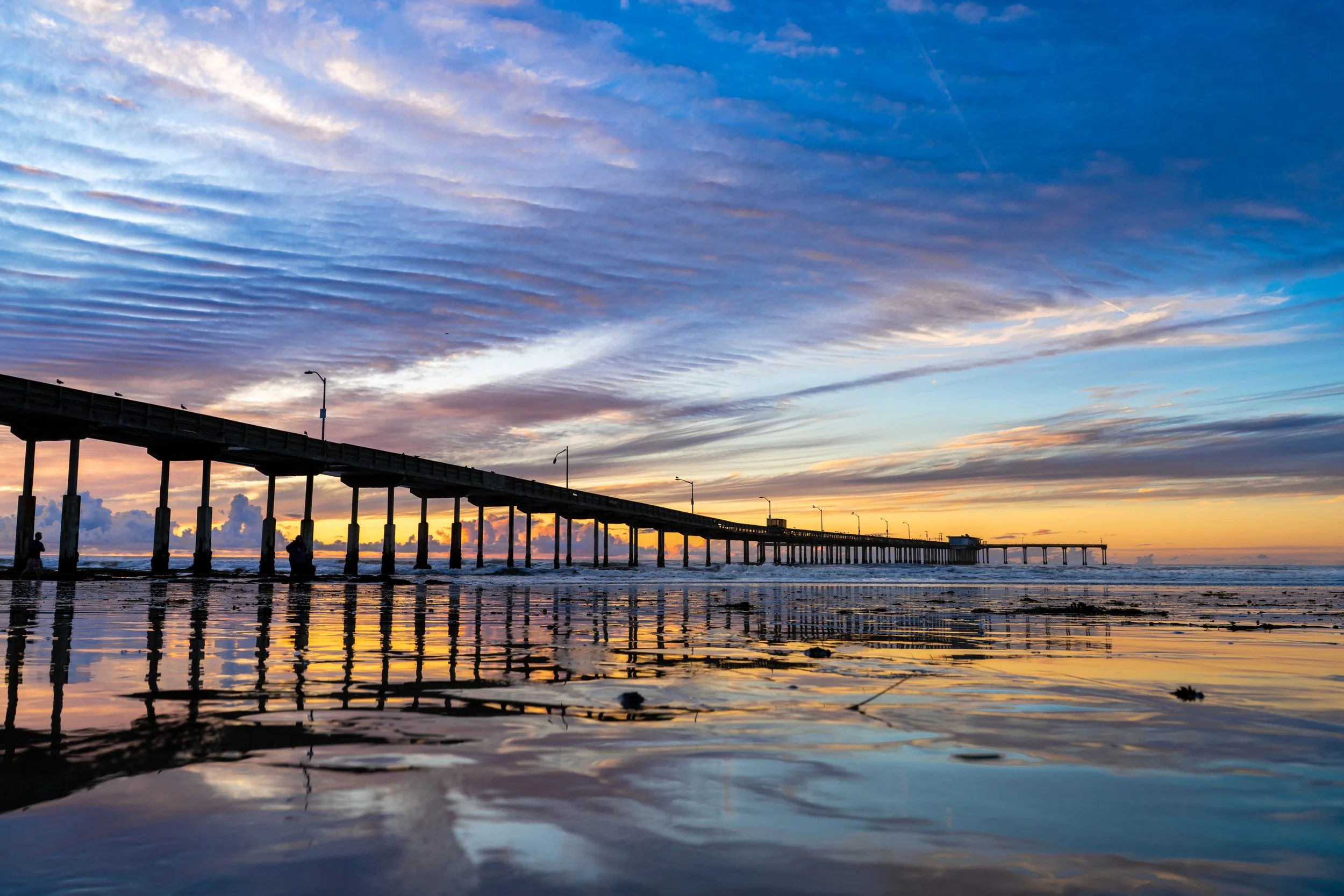 Sunset at a beach with a long pier extending into the ocean, reflecting colorful sky on shallow water.