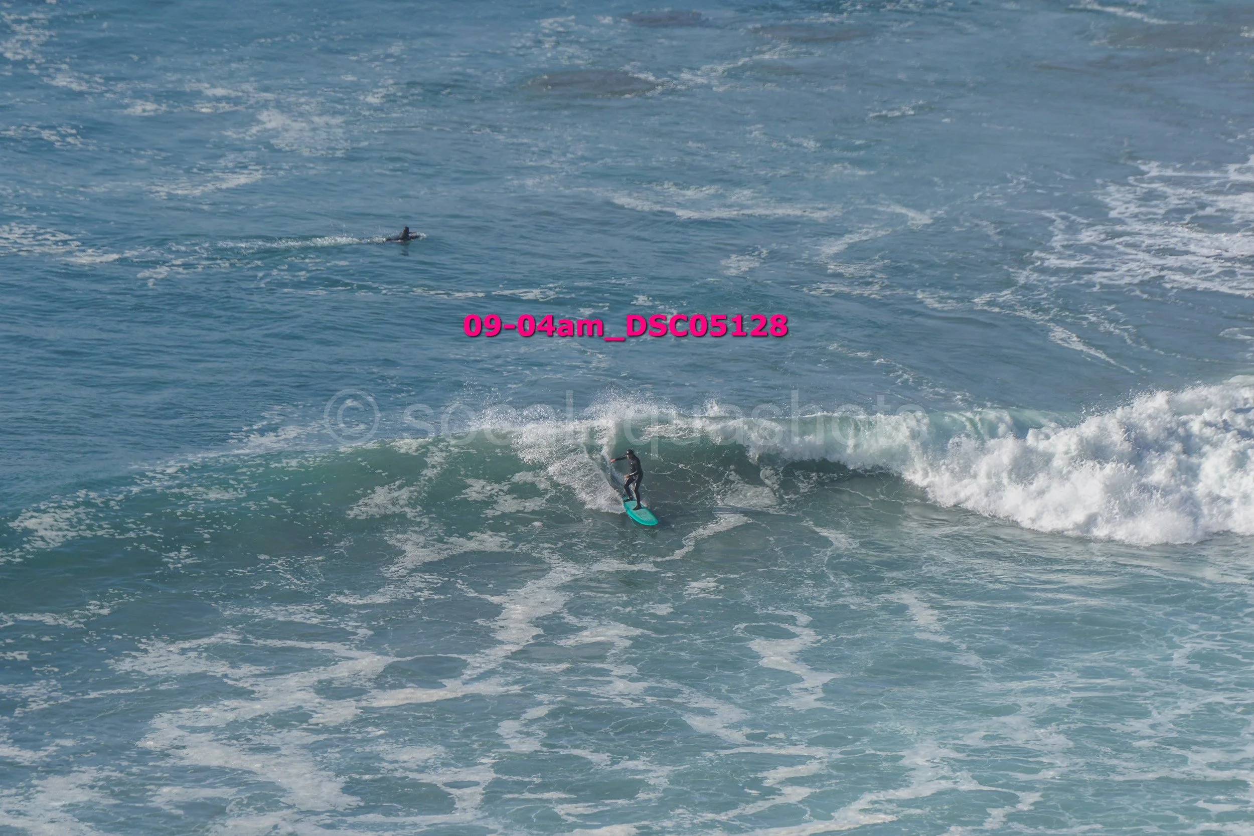 A person surfing on a blue surfboard in the ocean with waves around them. There is another smaller figure, possibly a swimmer or diver, visible in the water in the background.