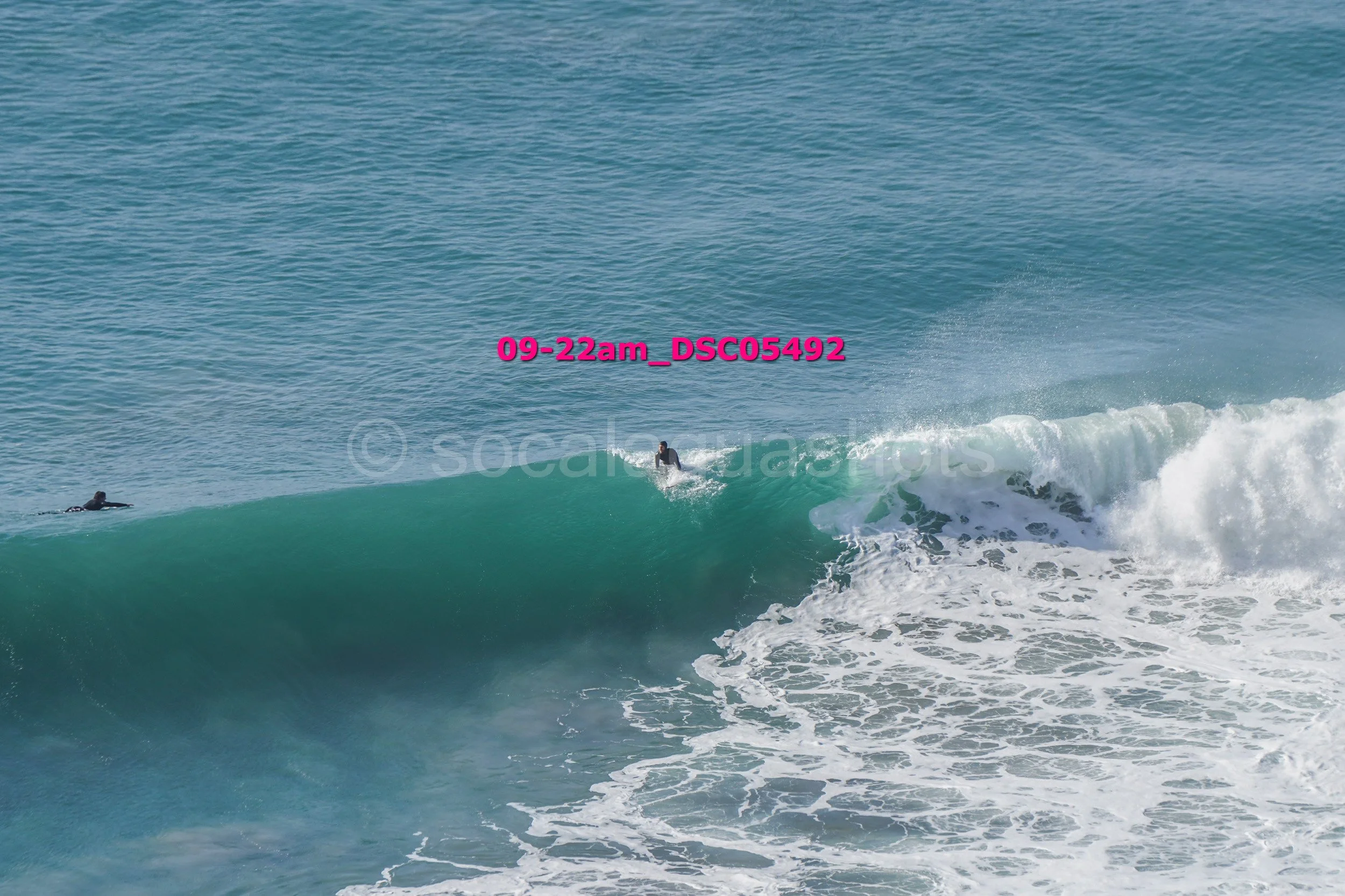 Surfer riding a wave in the ocean, with another surfer on a surfboard nearby.