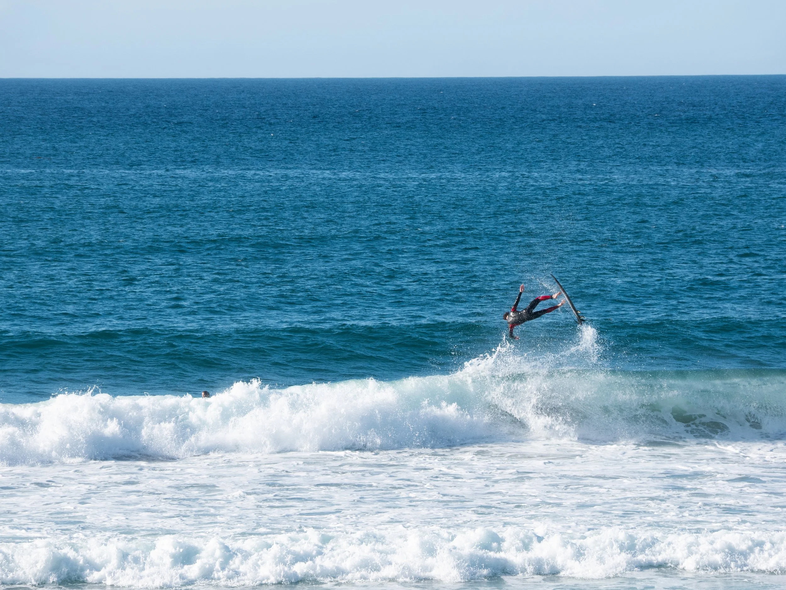 A person wearing a wetsuit performing an aerial stunt on a surfboard over the ocean waves with a blue sky background.