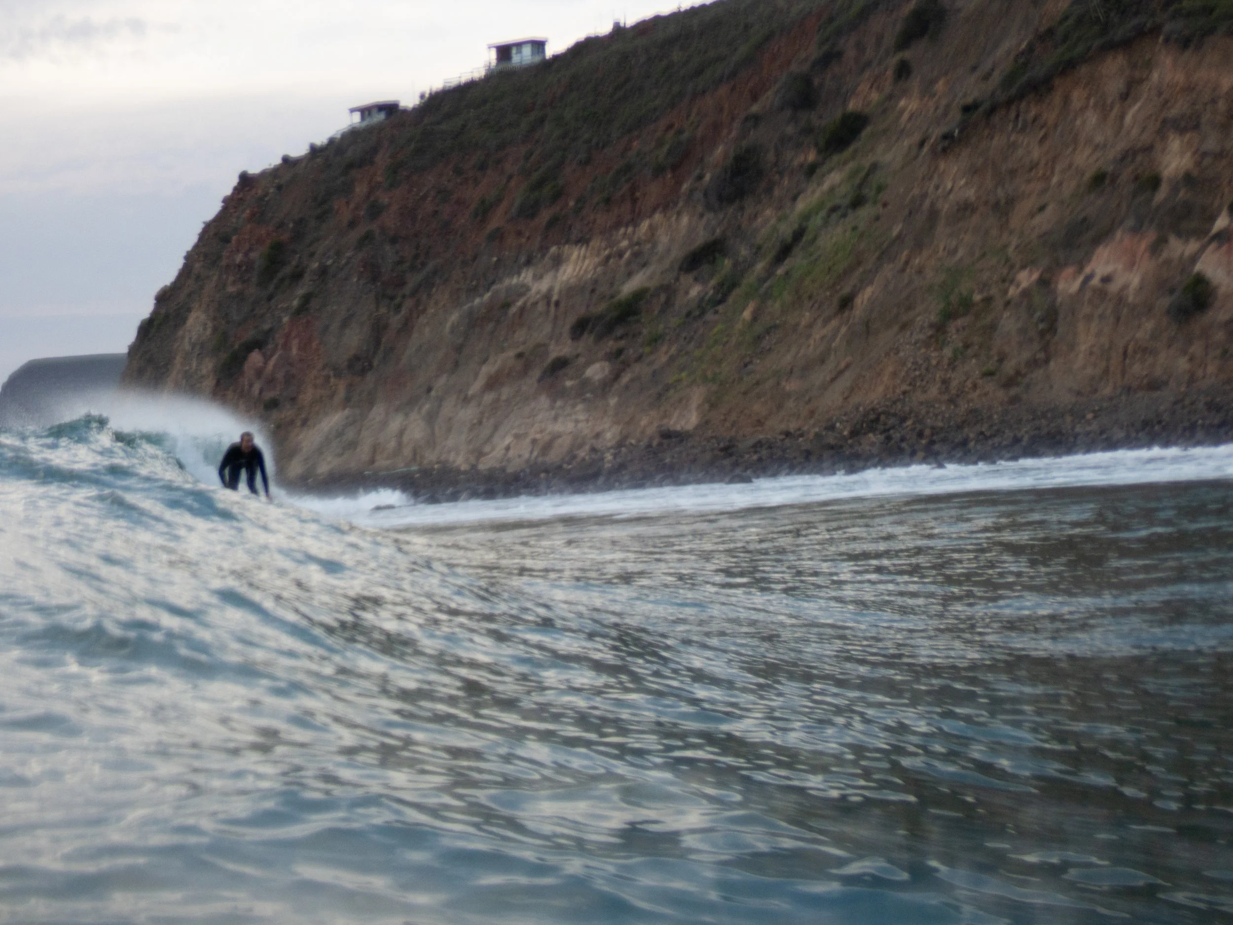 A person surfing on a wave near a rocky coastal cliff with houses on top during early evening or late afternoon.