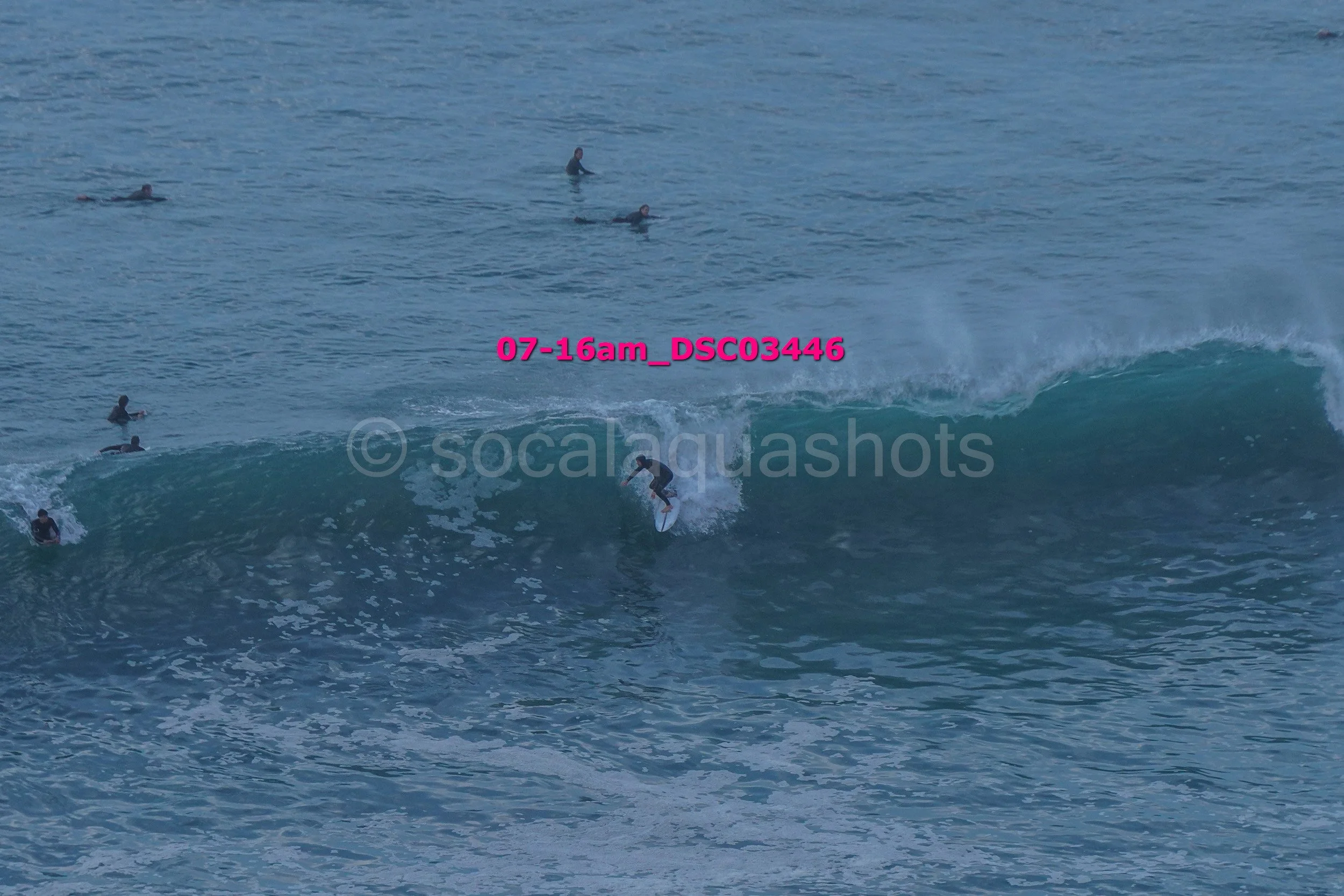 A person surfing on a wave with several surfers in the water around them, some waiting and some paddling, during daylight.