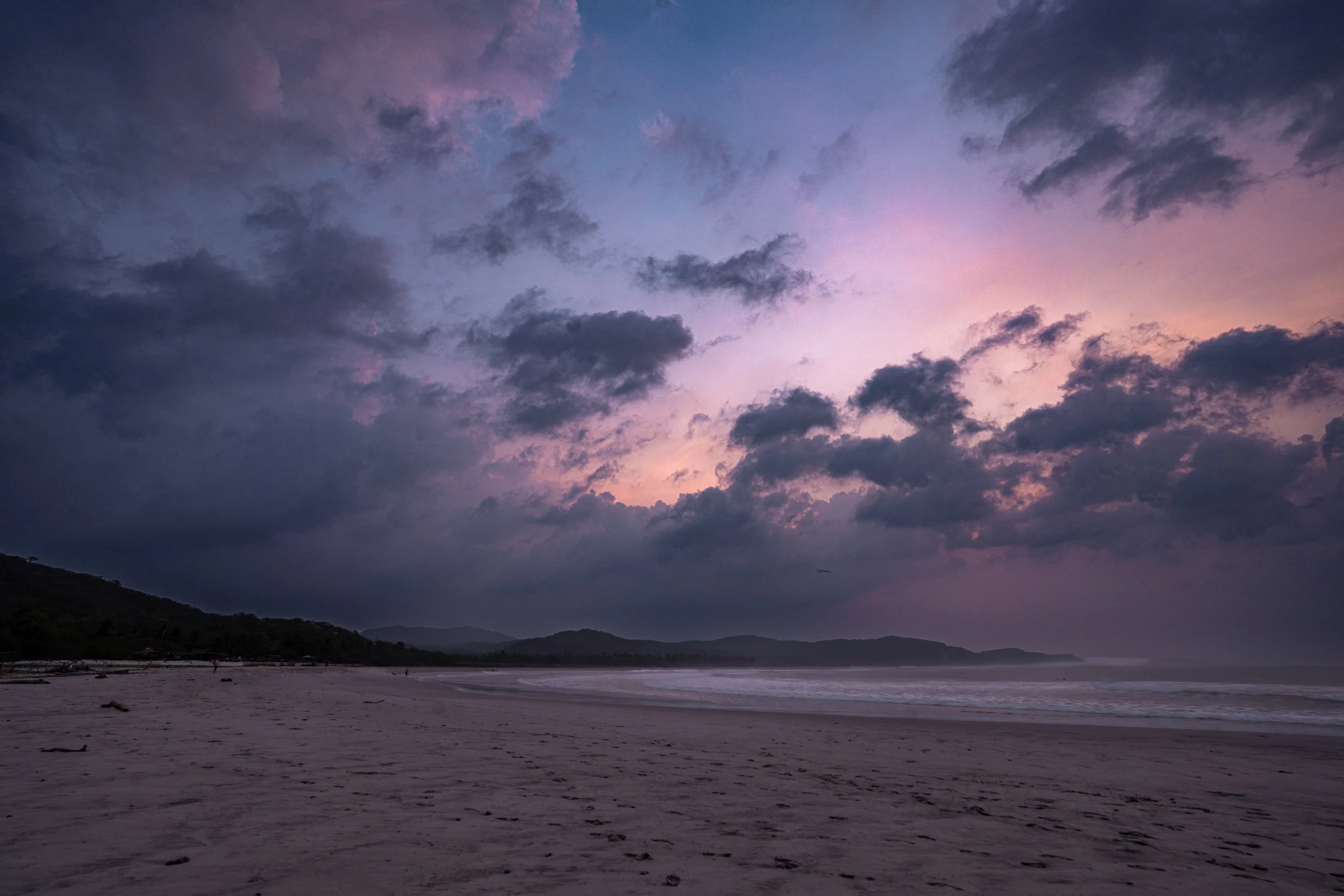 A beach at dusk with a cloudy sky showing shades of pink, purple, and blue, and mountains in the distance.