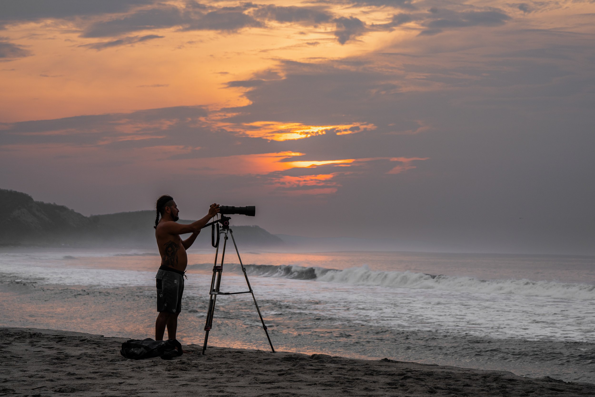 A man with long hair and tattoos stands on a beach at sunset, adjusting a camera on a tripod directed towards the ocean.