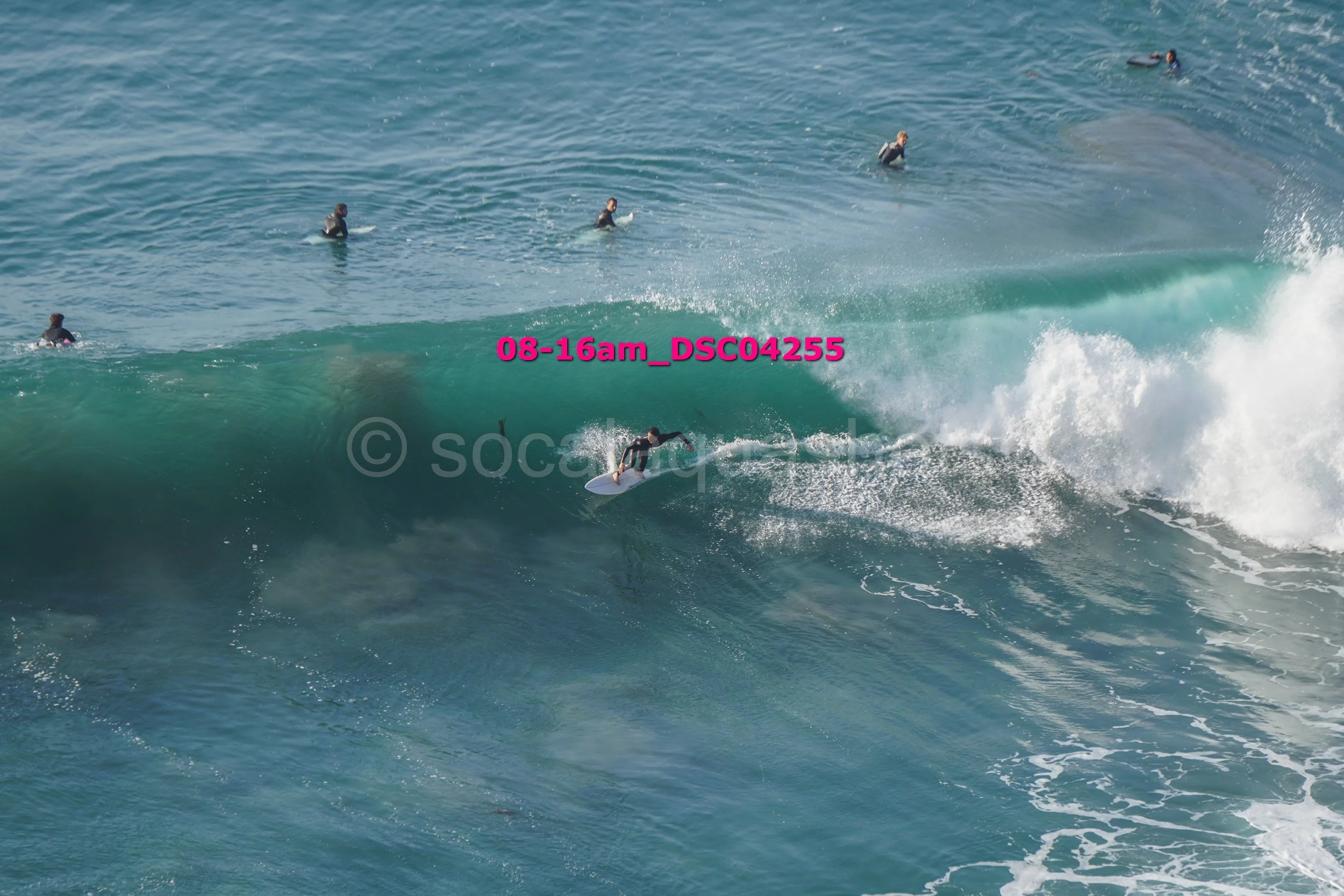 A person surfing on a large wave in the ocean, with several other surfers floating in the water nearby.