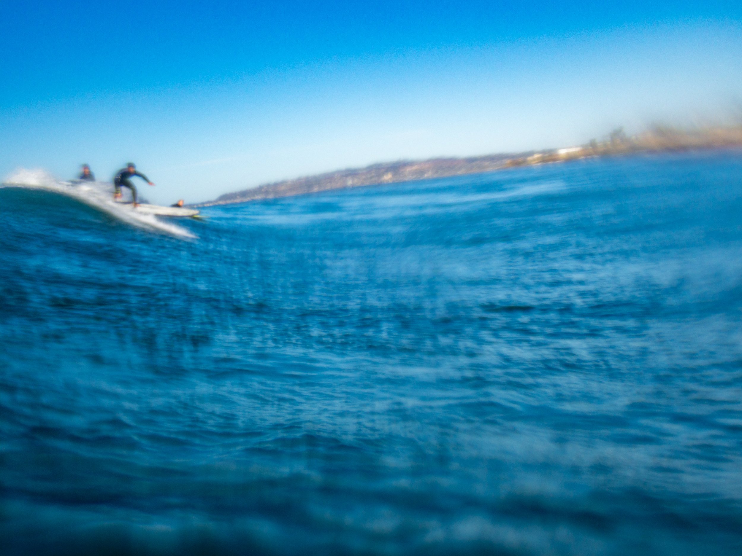 Two surfers riding a wave in the ocean under a clear blue sky with distant shoreline.