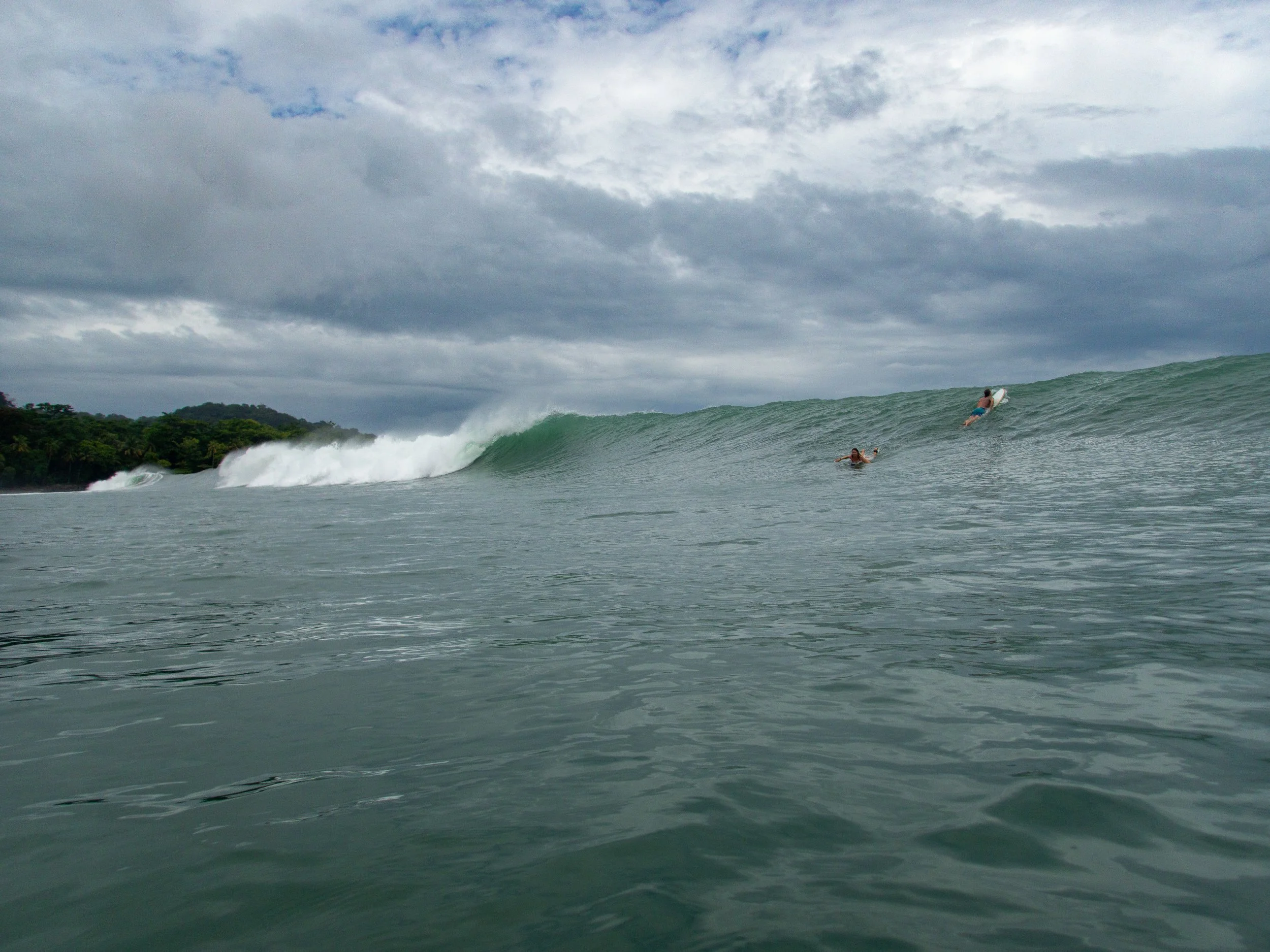 Surfers paddling toward a large ocean wave under a cloudy sky.