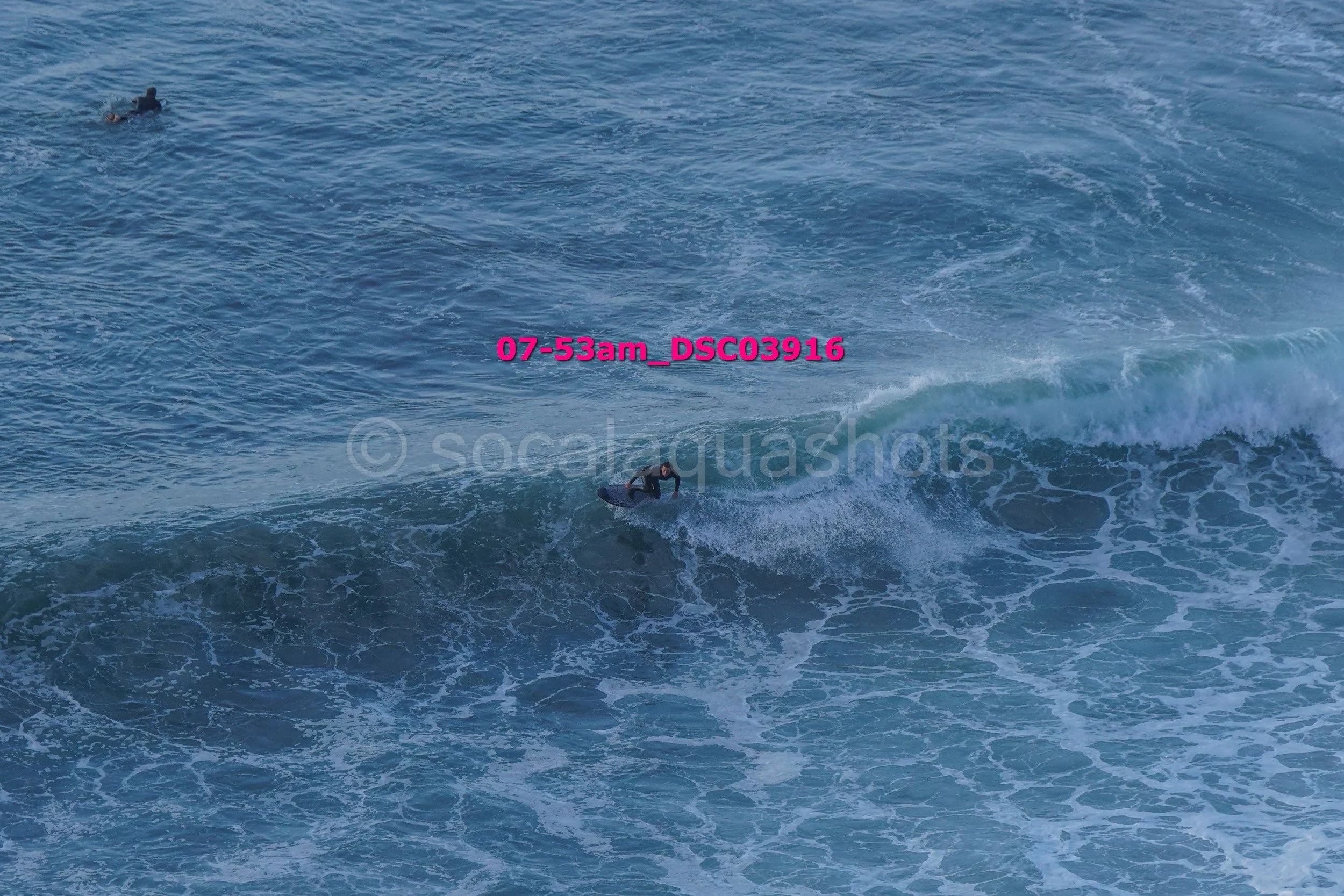 A person surfing on a wave in the ocean during daytime, with another person swimming in the water nearby.