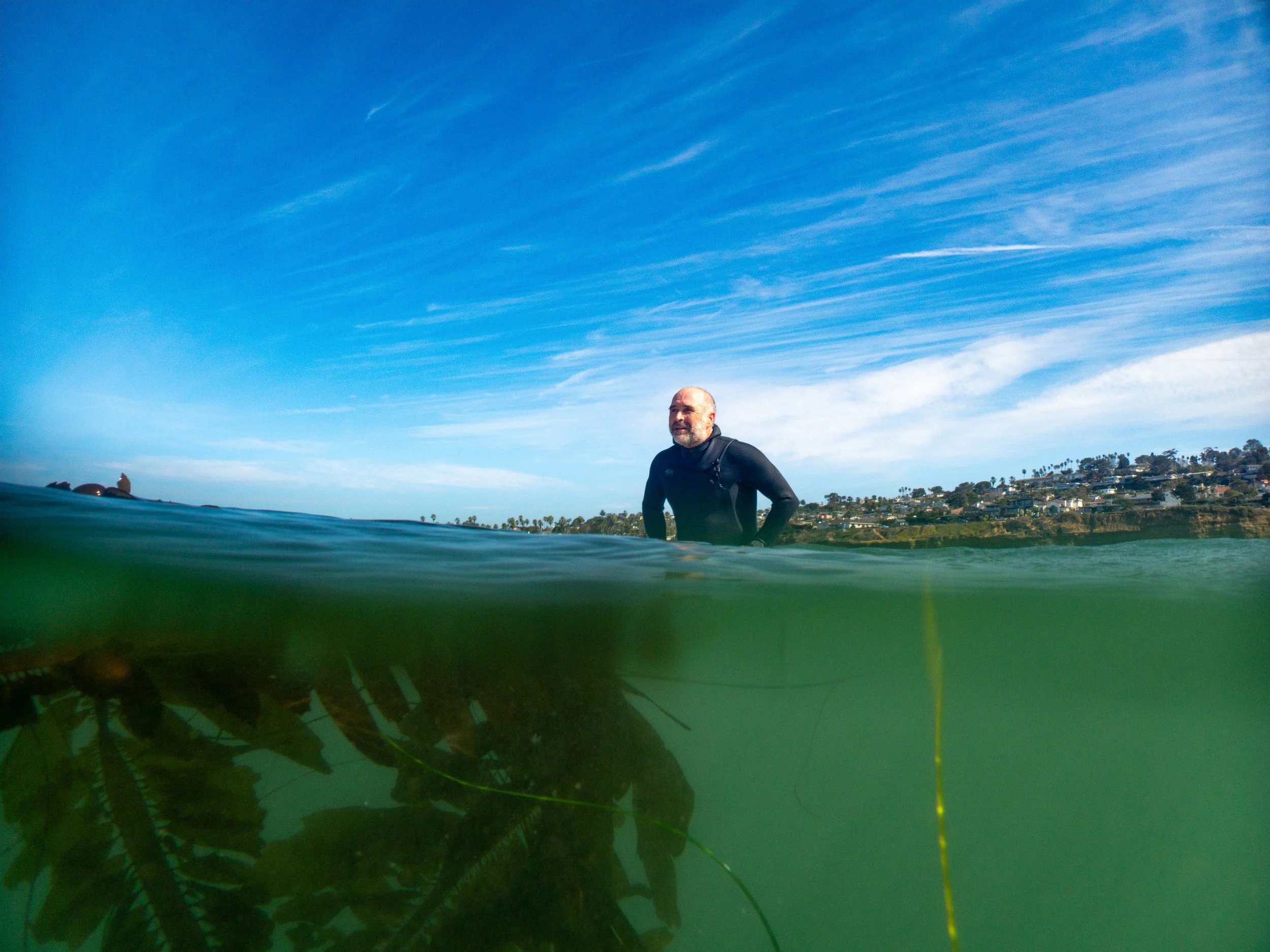 A man wearing a wetsuit standing in the water near a coastline with houses and palm trees, with a blue sky and wispy clouds above.