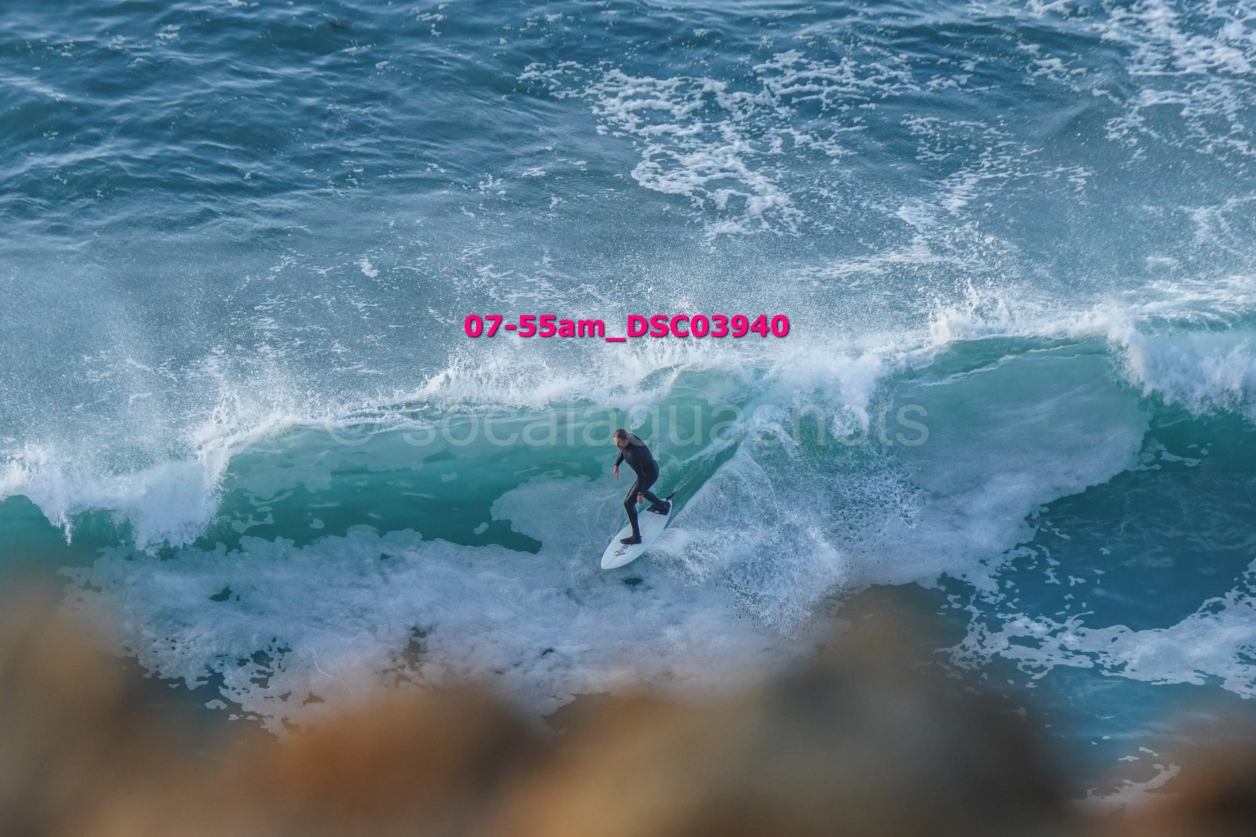 A person surfing on a wave in the ocean with a rocky foreground.