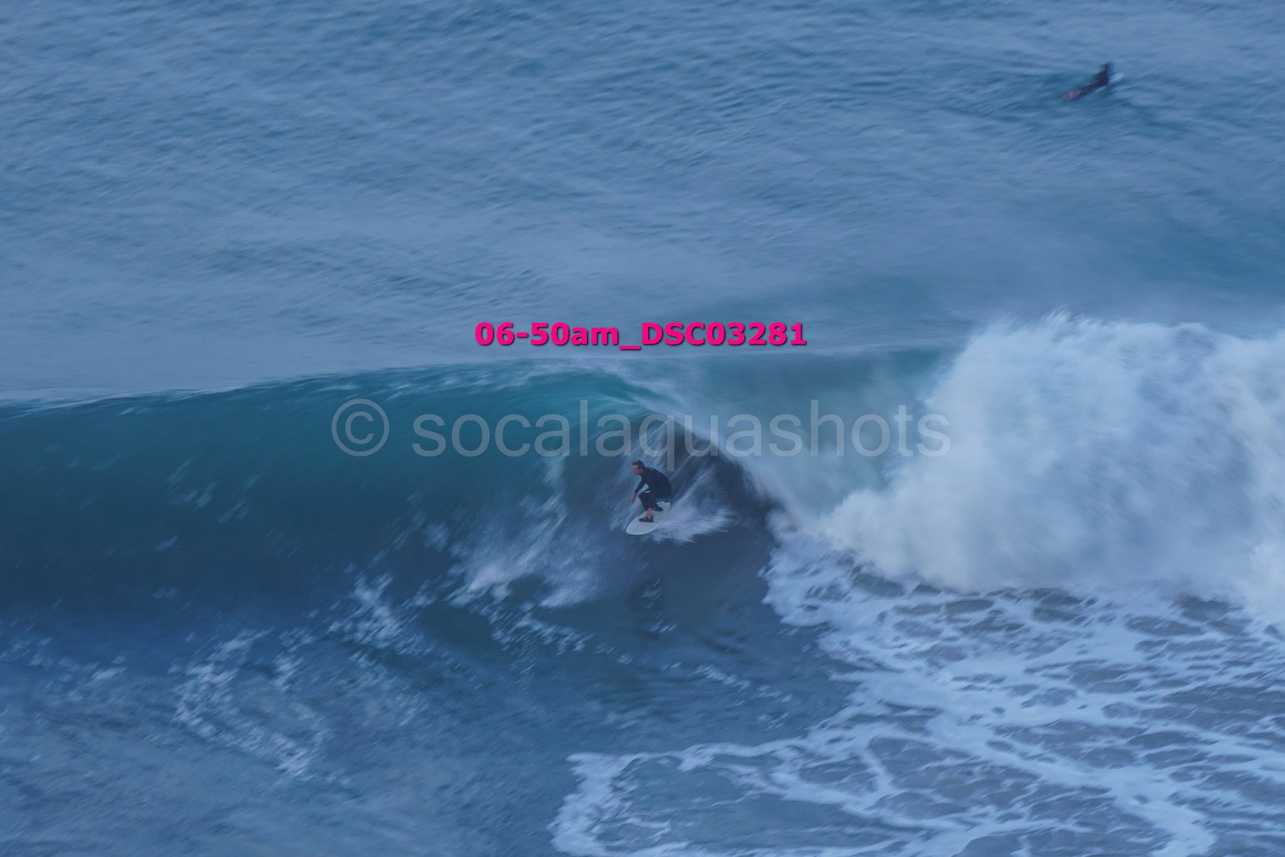A person surfing on a wave in the ocean with another surfboarder visible in the water in the background.