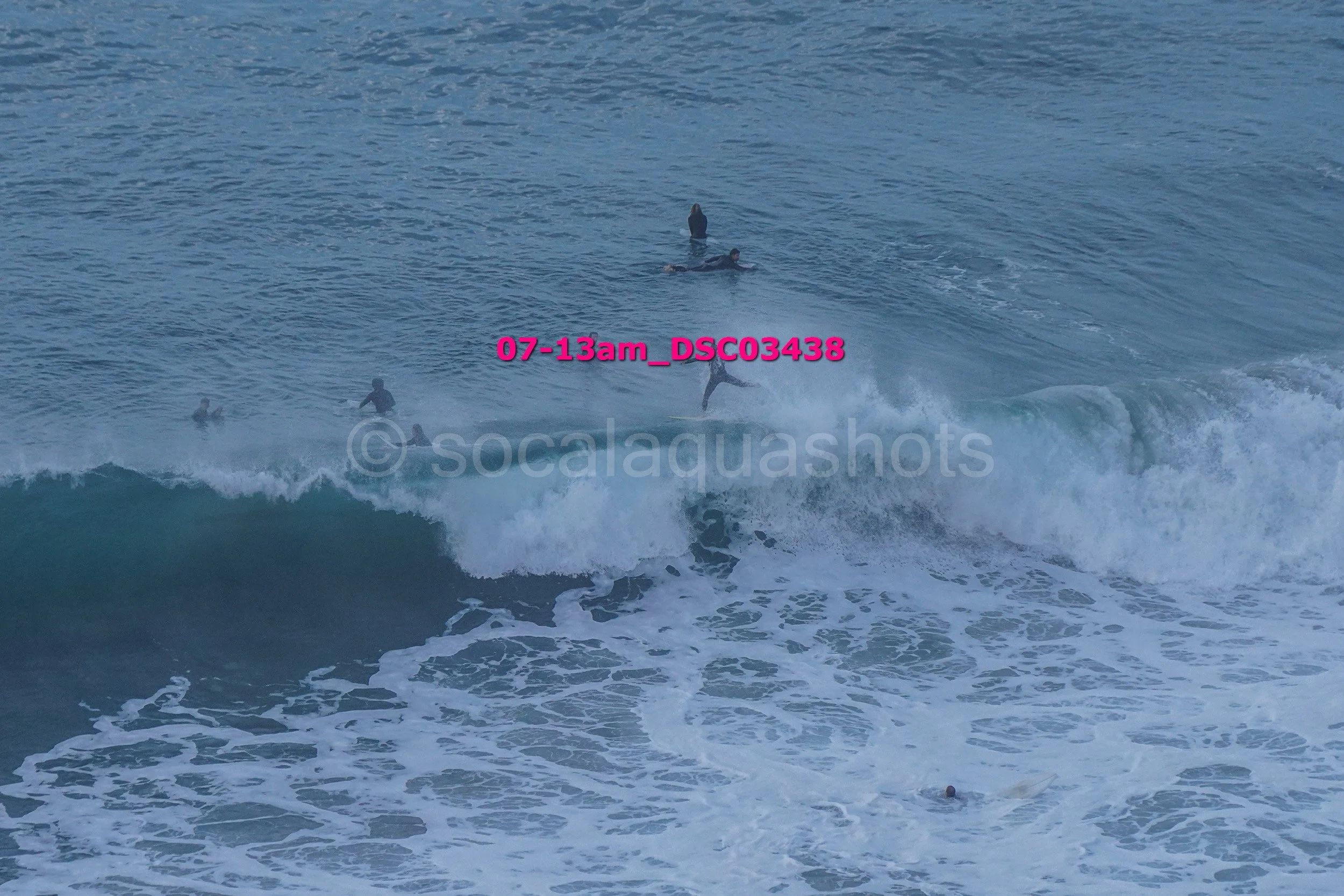Surfers in the ocean waiting for waves, with one surfer riding a wave and others floating or paddling nearby.