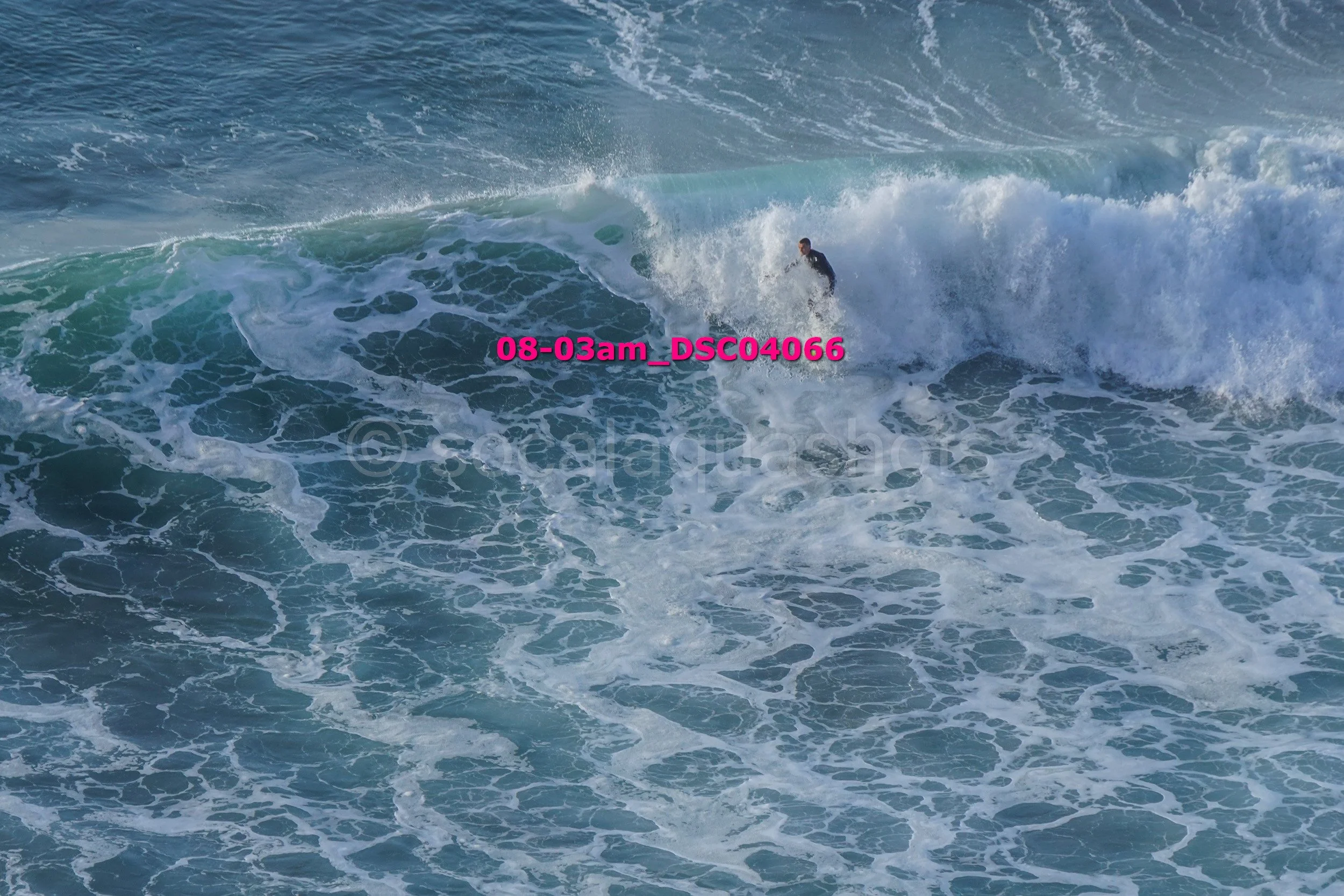 A person surfing on a large ocean wave with white foam, water spraying, and the sky visible in the distance.
