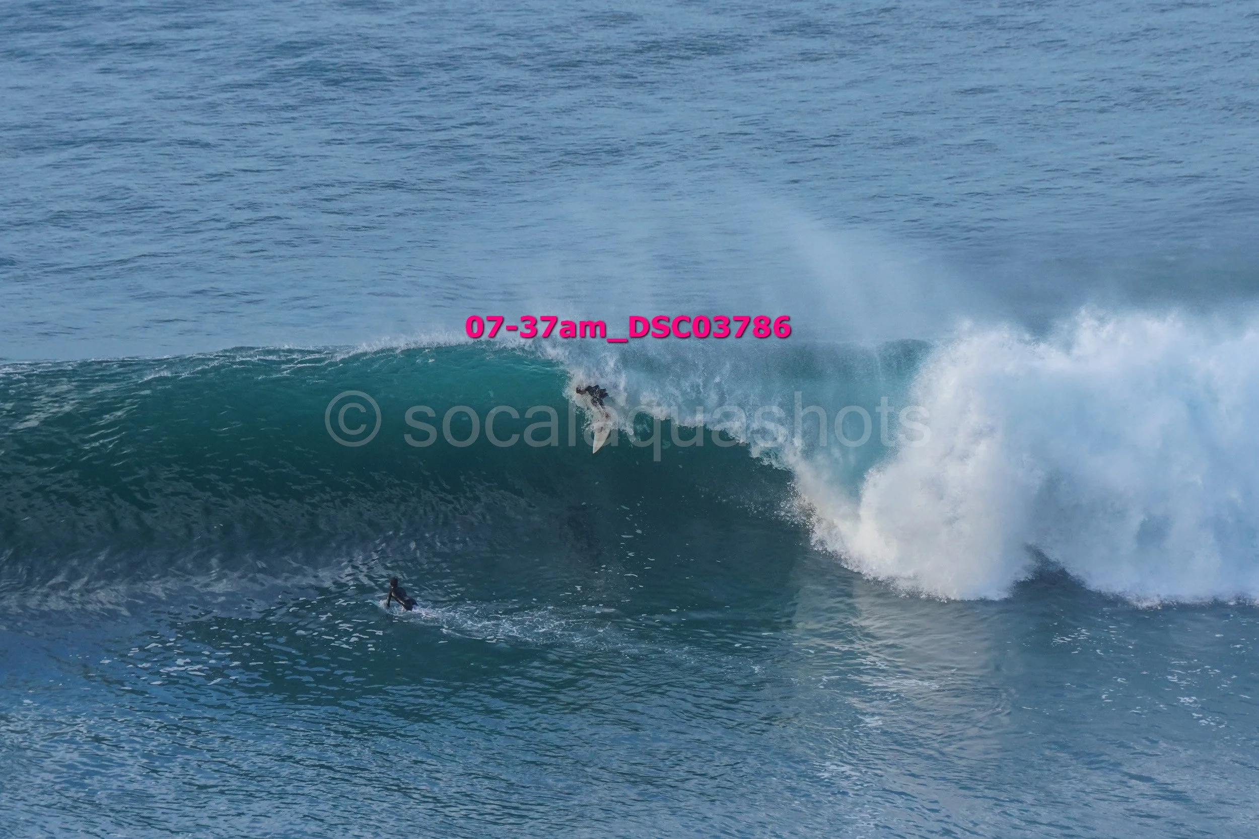 A surfer riding inside a large ocean wave with another surfer paddling below.