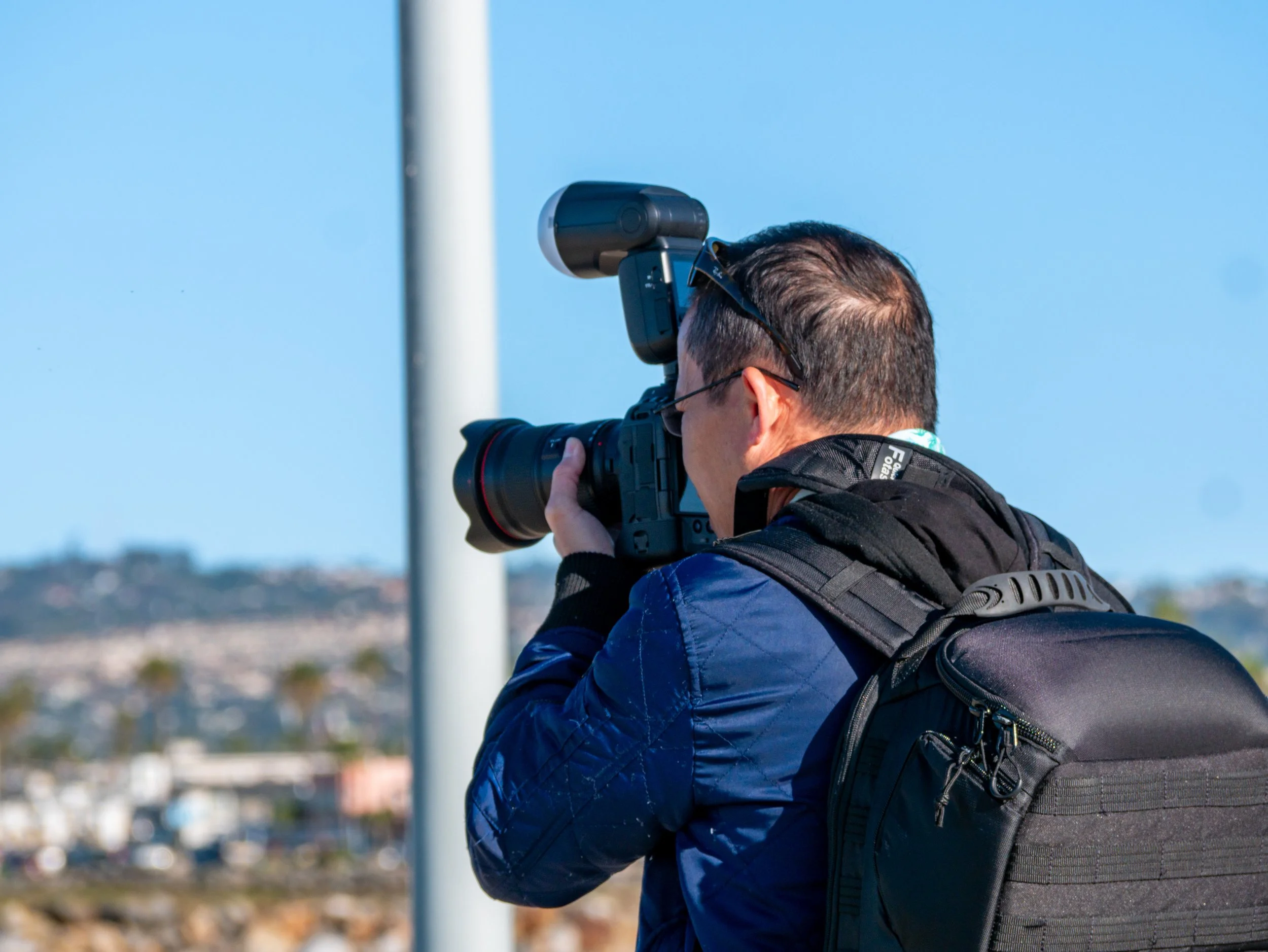 A man with a backpack and sunglasses taking a photo with a camera in an outdoor setting against a blue sky.