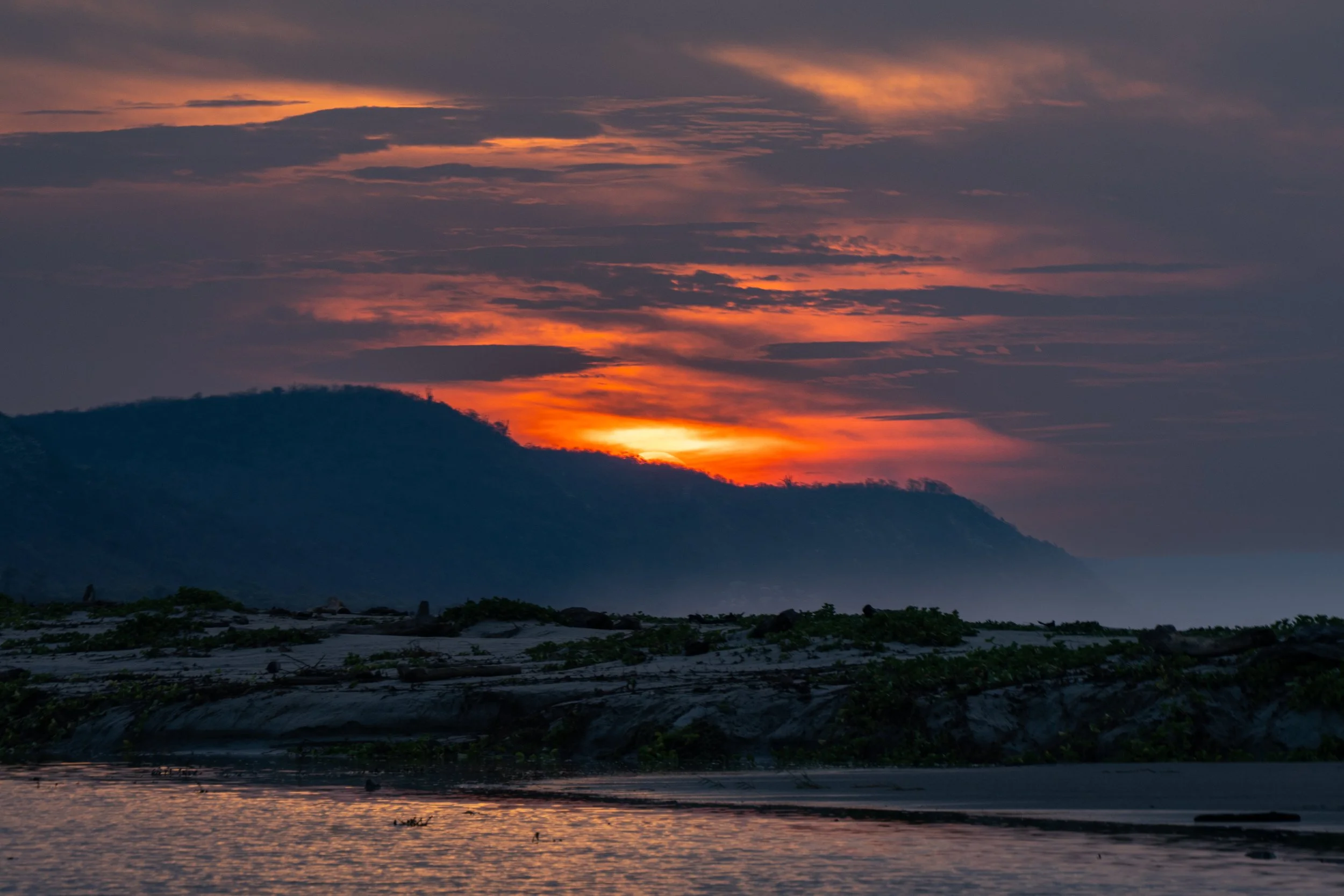 A sunset over a mountainous landscape with a river in the foreground, with partly cloudy skies and vibrant orange and purple hues.