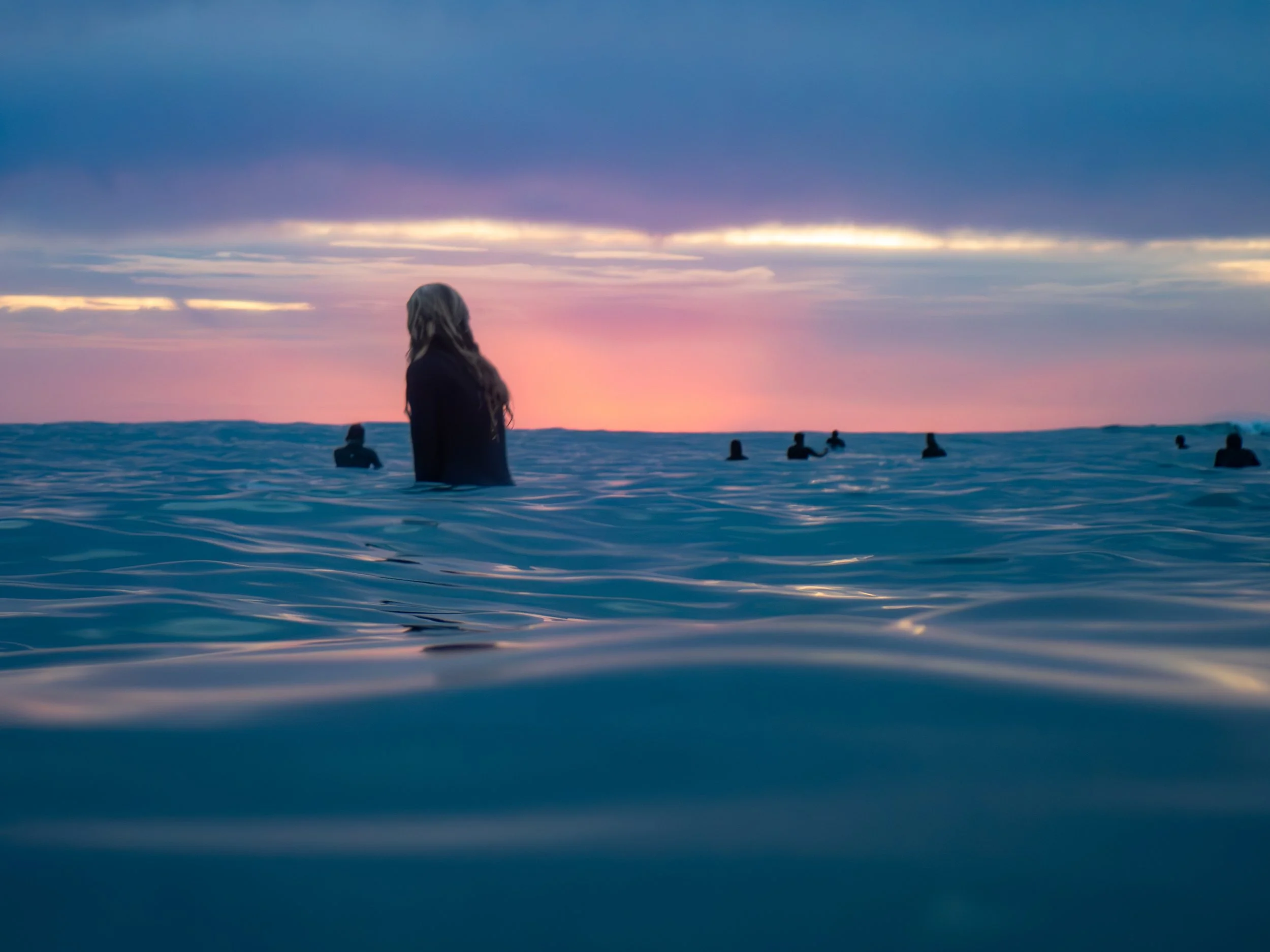 People swimming in the ocean during sunset with colorful sky and calm blue water.