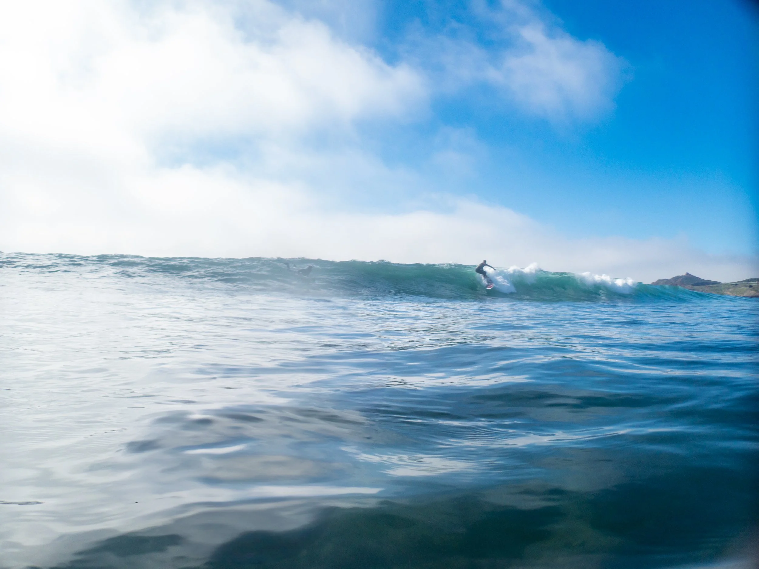 A person surfing on a wave in the ocean with a blue sky and some clouds overhead.
