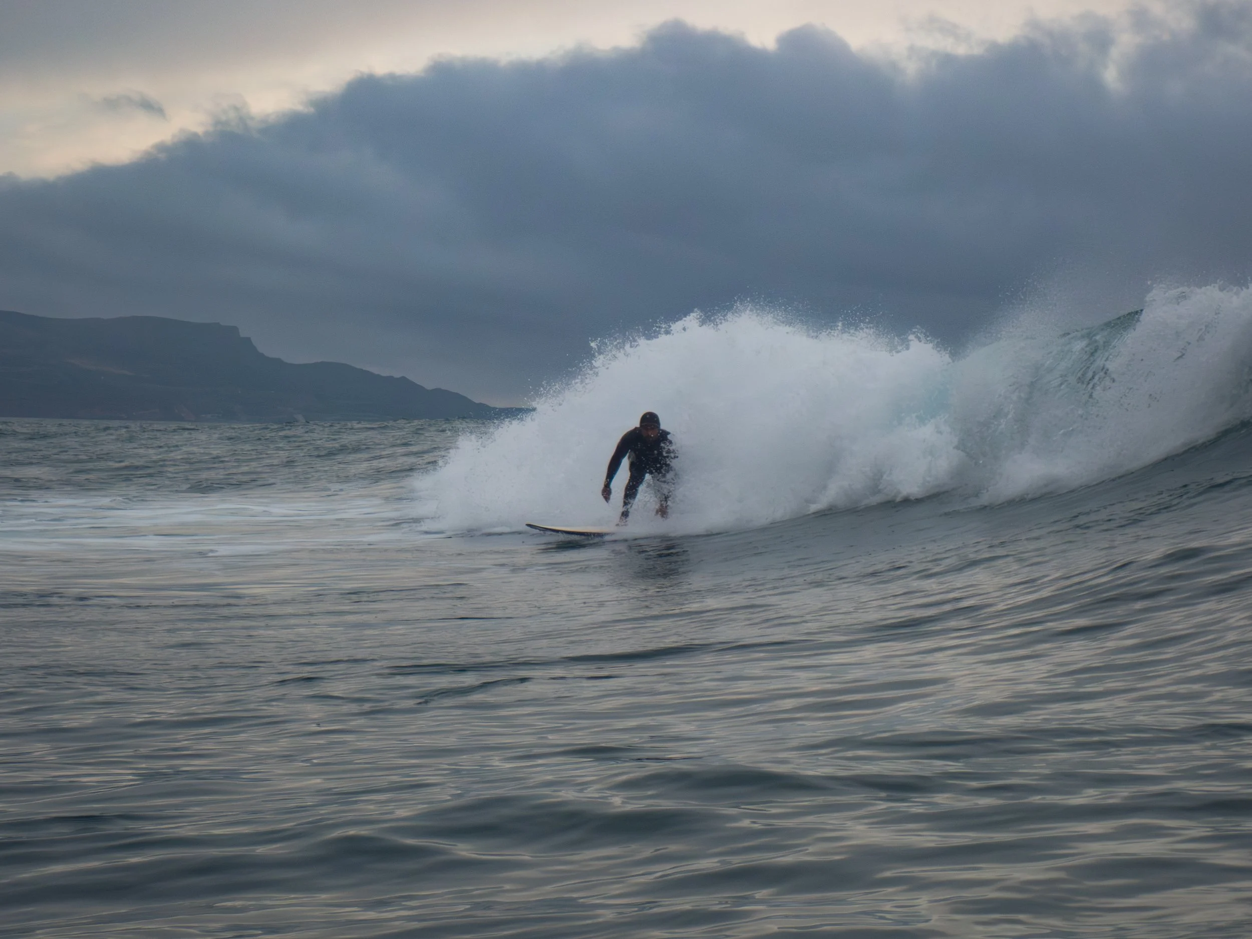 A person surfing on a large wave in the ocean with a cloudy sky and distant mountains in the background.