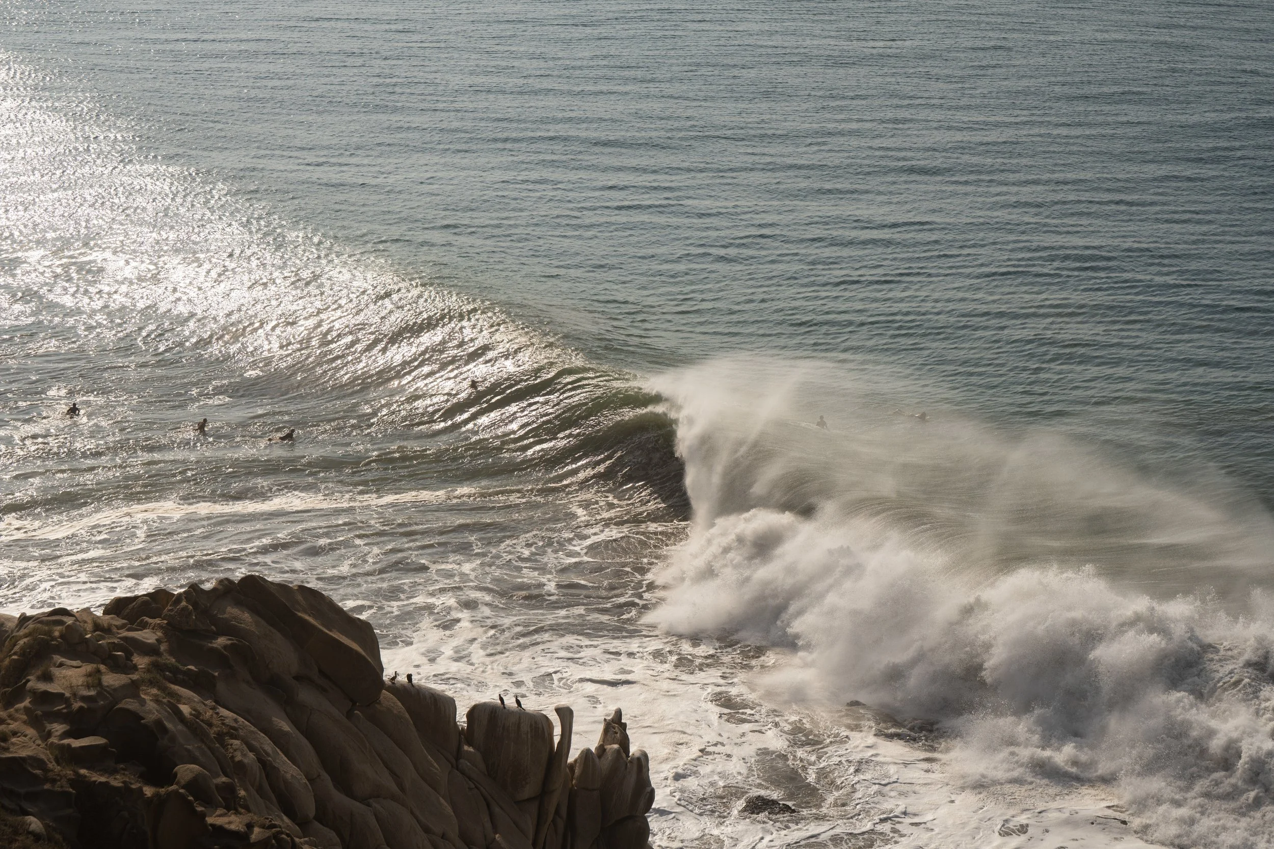 Ocean waves crashing against rocks with surfers in the water at sunset.