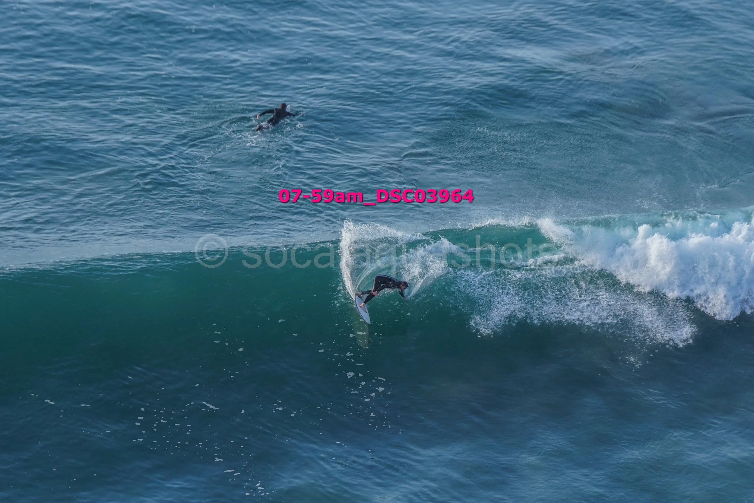 A person in a black wetsuit surfing on a wave with another person swimming nearby in the ocean.