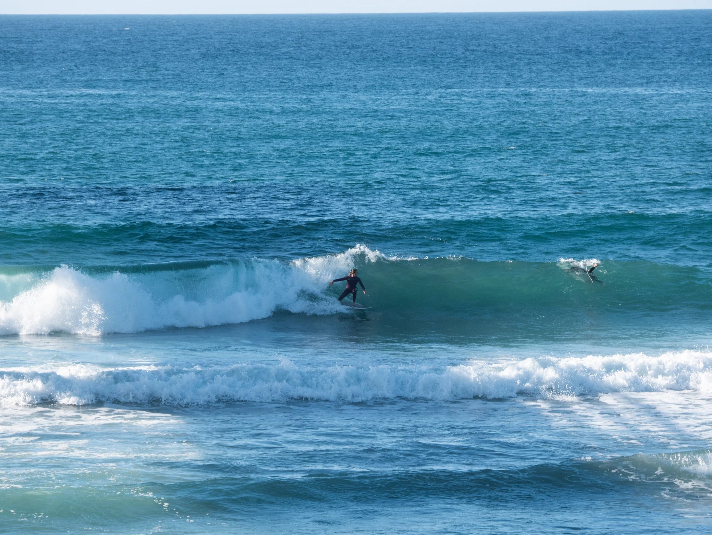 A person surfing on a wave in the ocean, with another person paddling on a surfboard nearby.