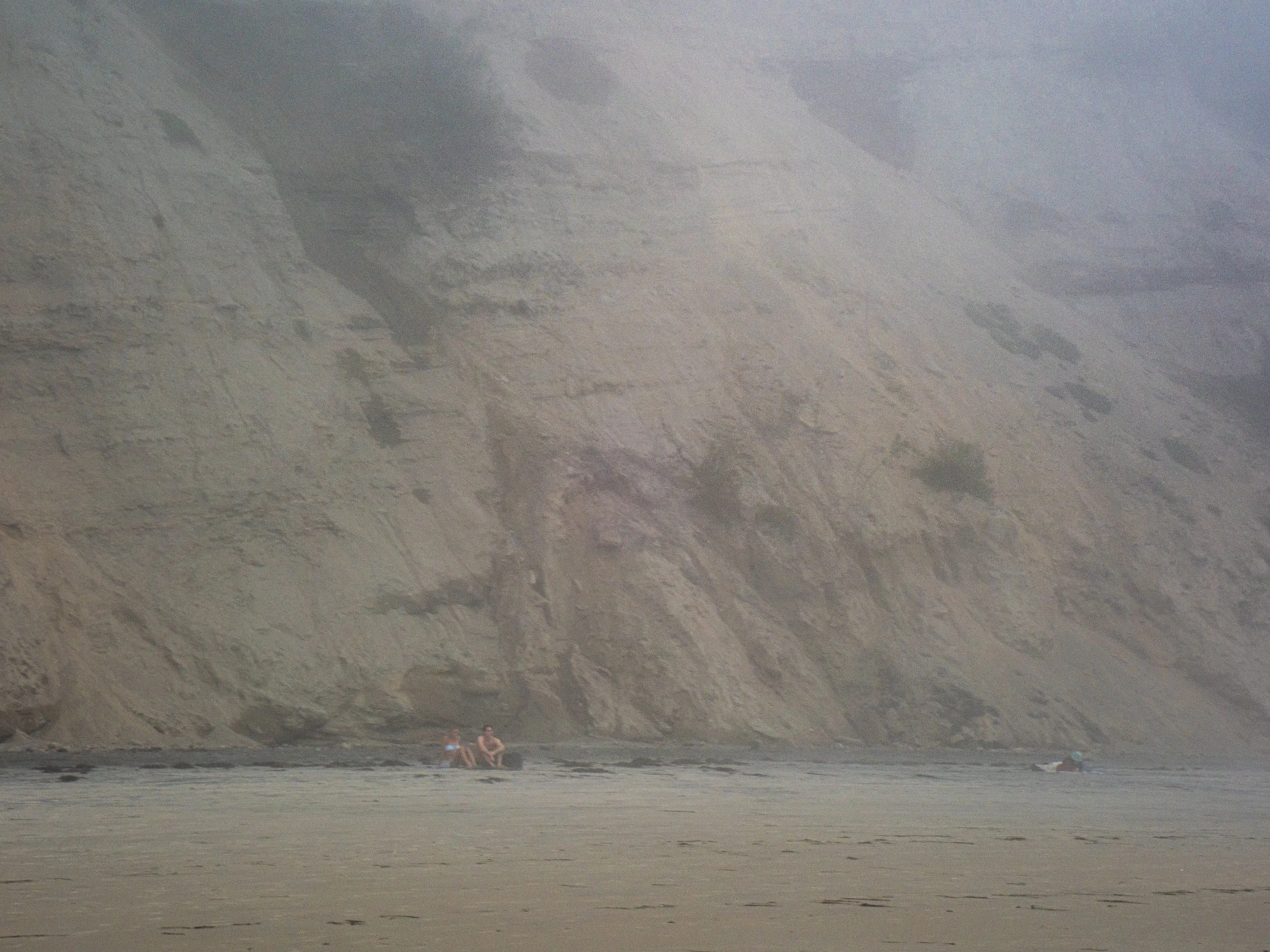 People sitting on a beach near a large rocky cliff with some sand and a hazy sky.