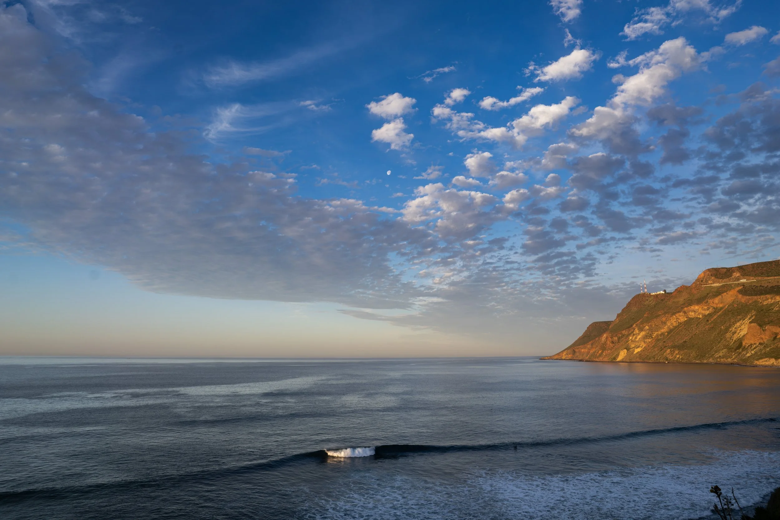 A scenic coastline with a cliff on the right side, the ocean in the foreground, and a sky filled with scattered clouds and a visible moon.