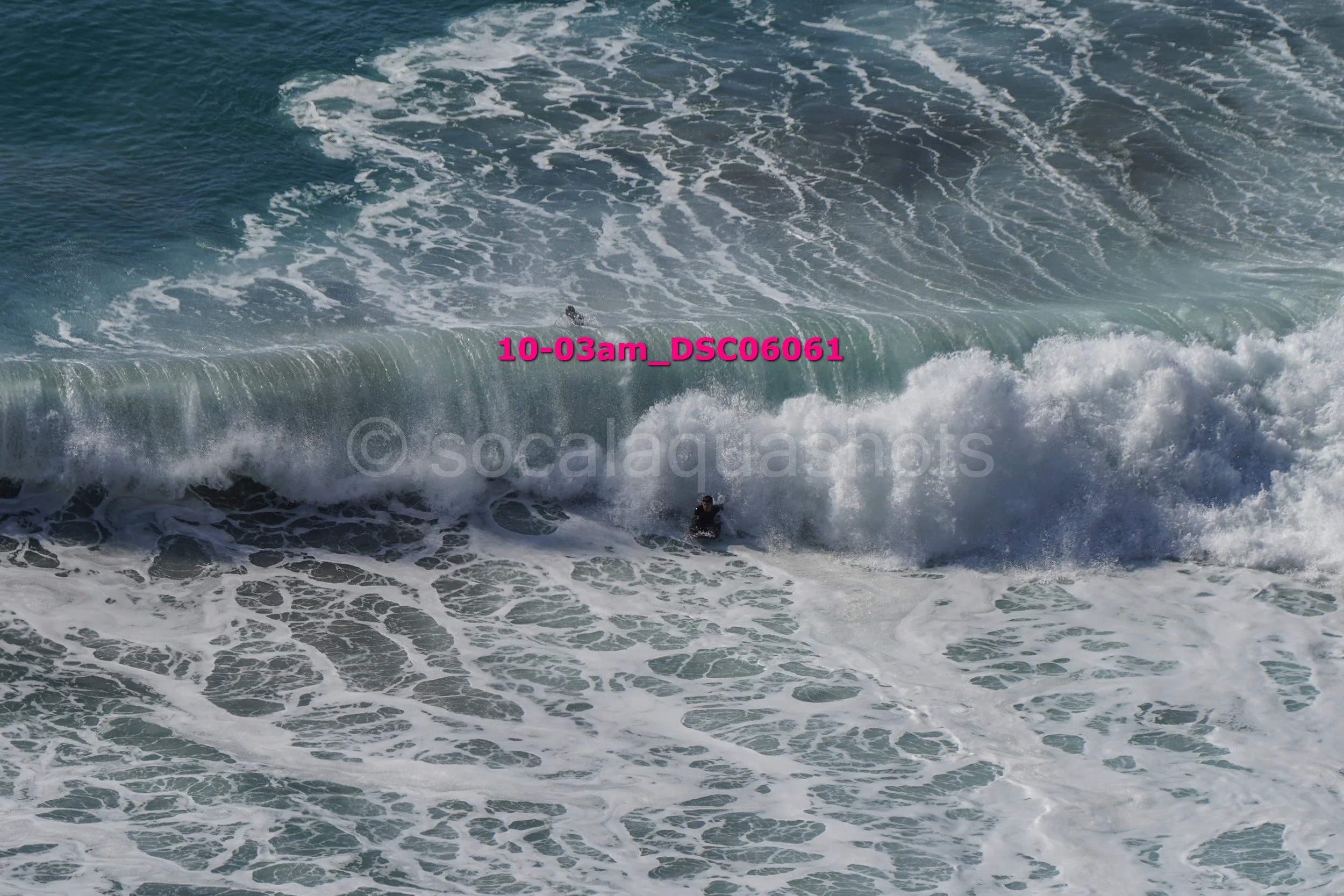 Large ocean wave with a surfer underneath, partially obscured by white foam, with a pink watermark text across the wave.