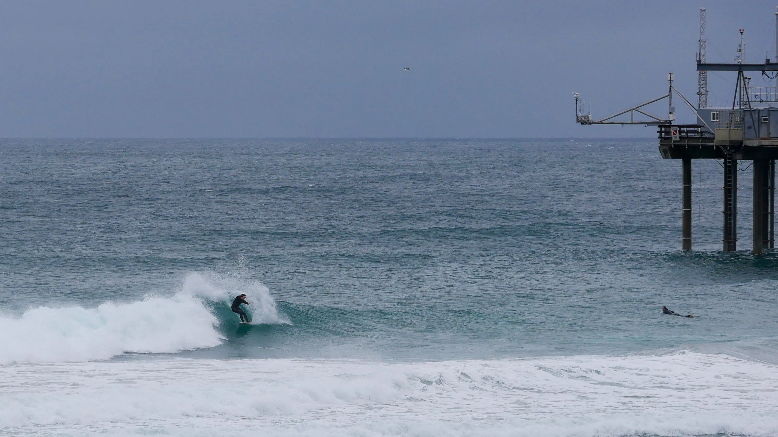 Surfer riding a wave near a pier with an overcast sky and ocean in the background.