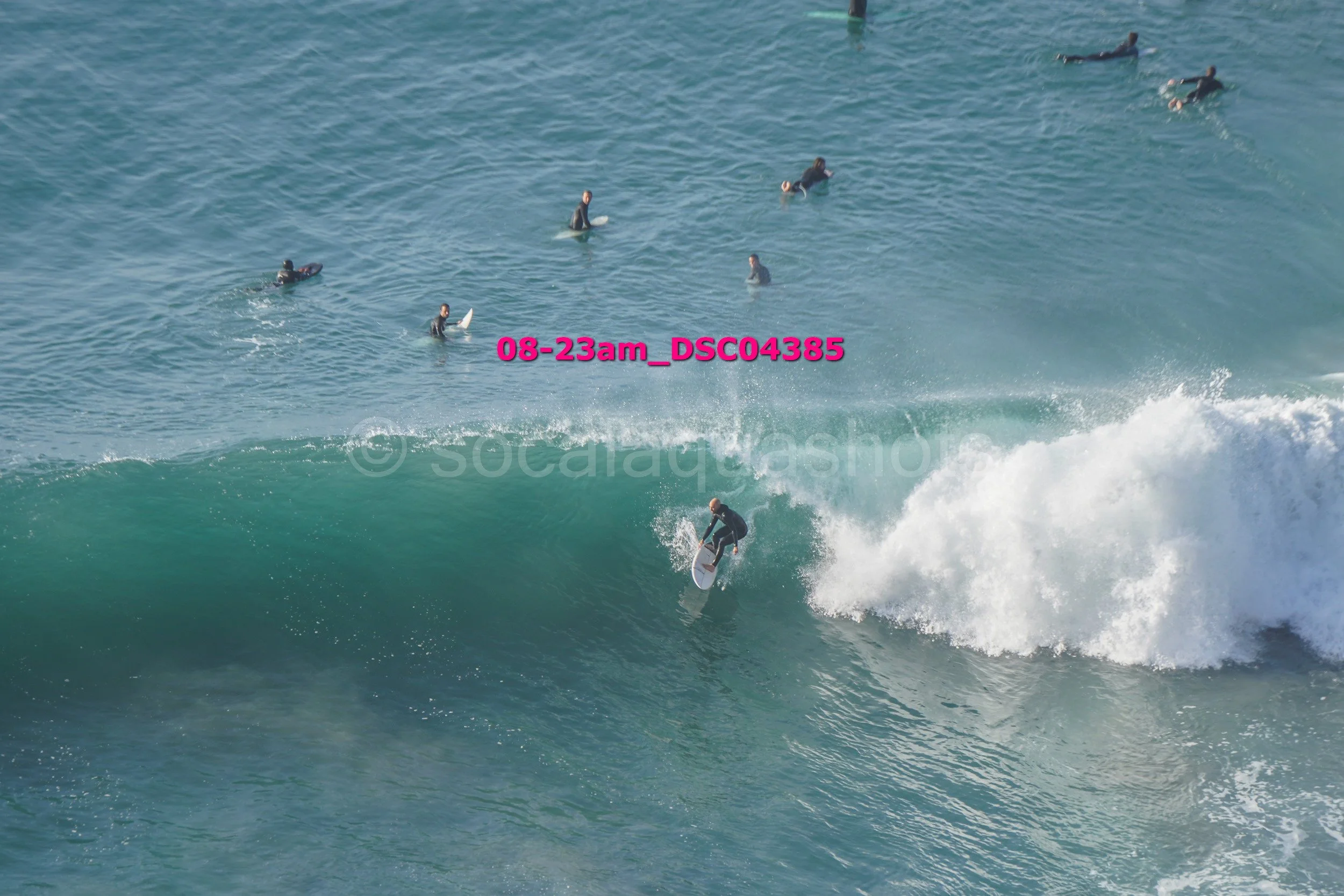 Surfer riding a large wave while several surfers wait in the water behind him.