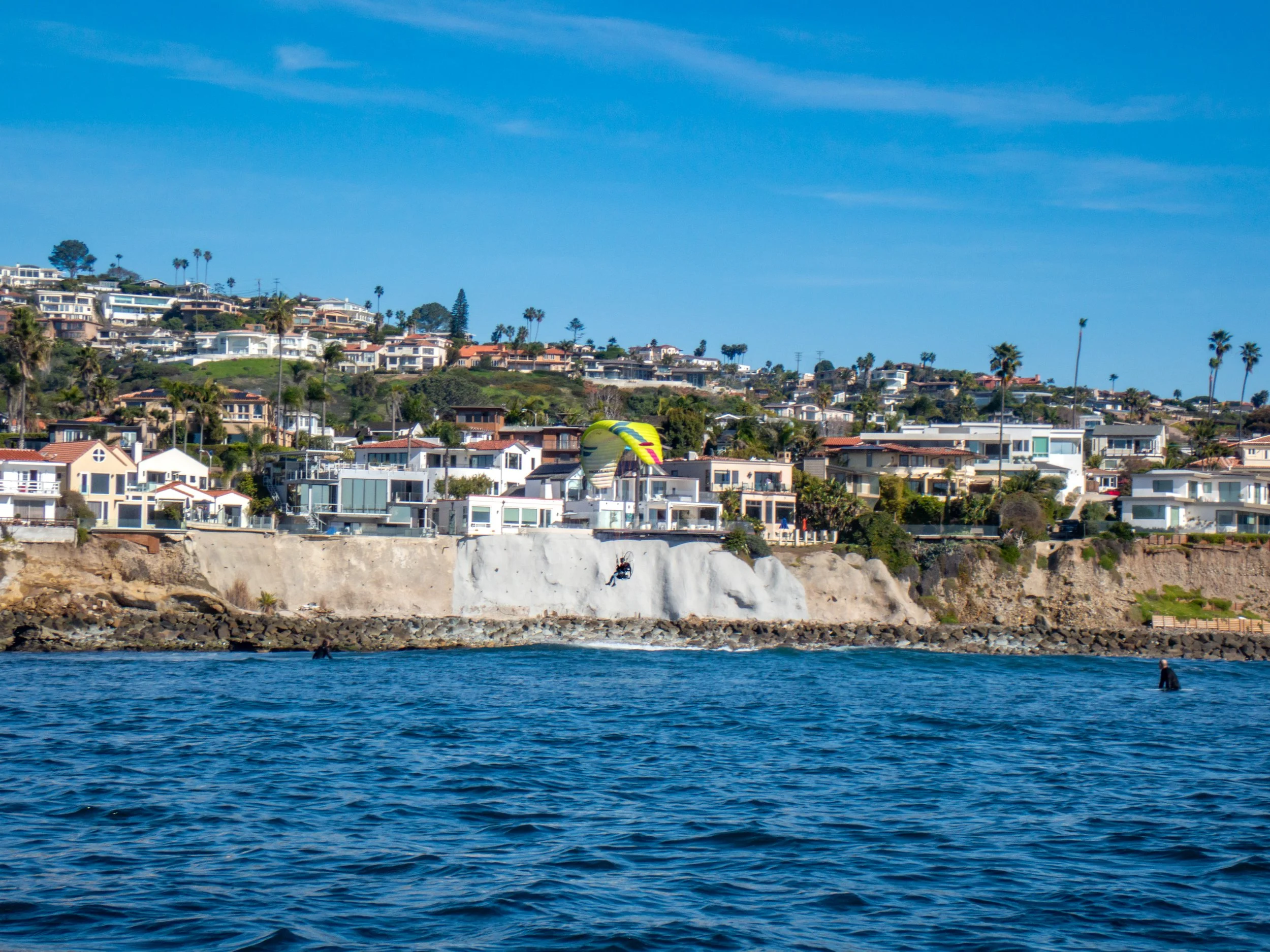 A coastal scene with houses on a hillside, a paraglider in mid-air over the water, and people in the water near the shore on a sunny day.