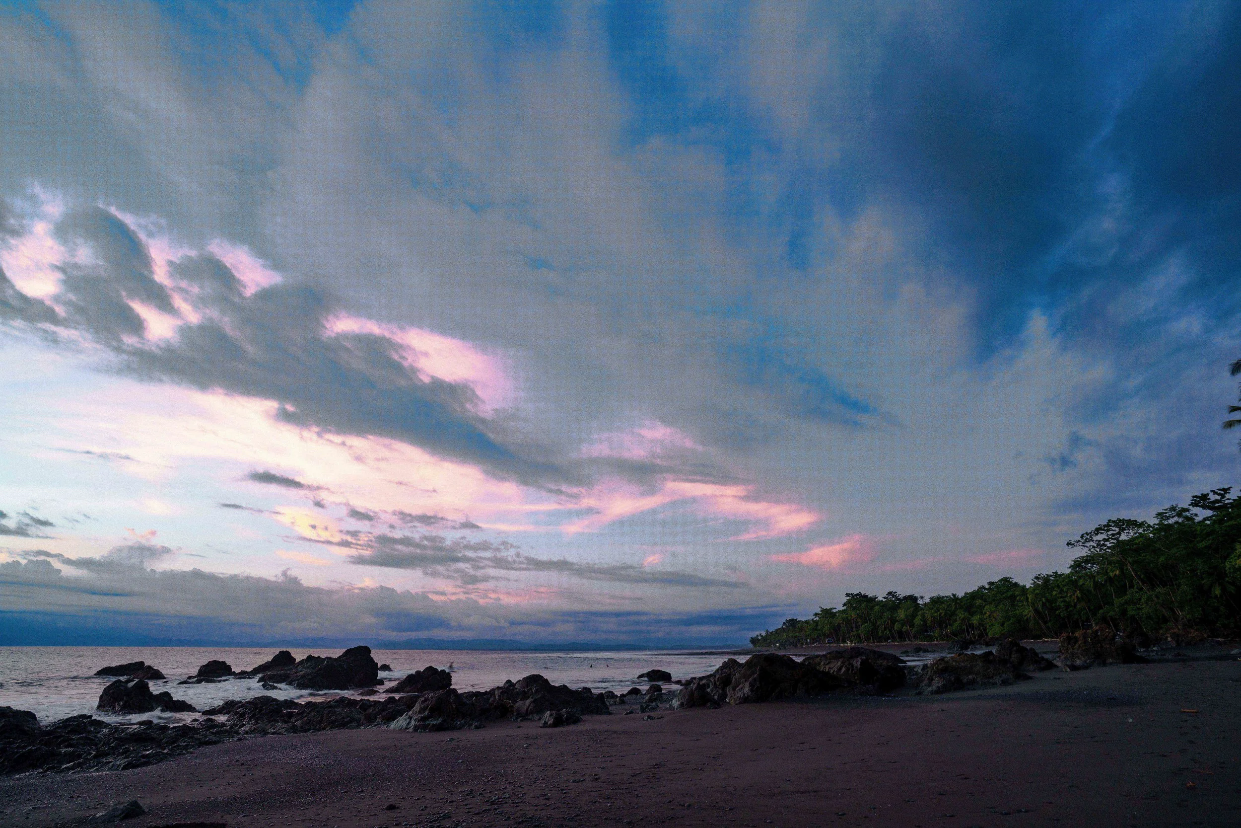 A coastal landscape during sunset with a cloudy sky, rocky shoreline, and silhouette of trees on the right.