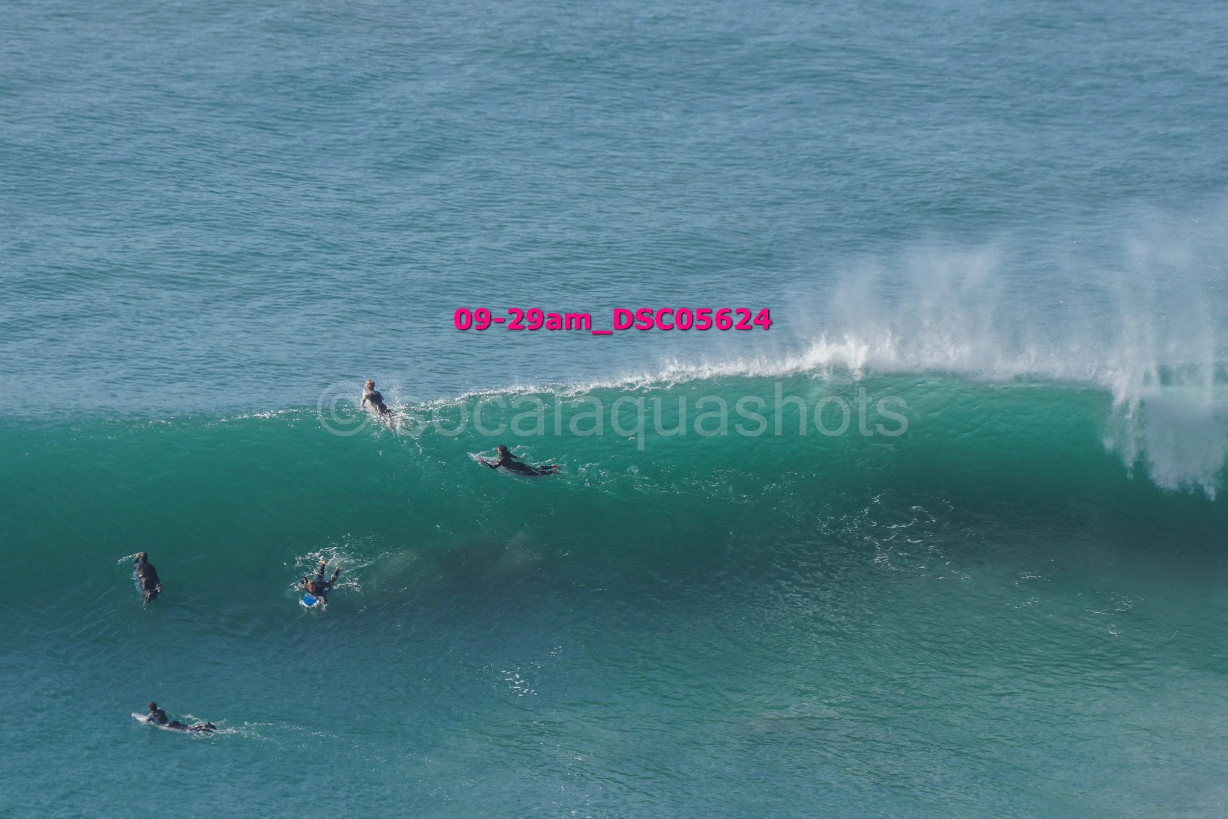 Surfers riding and paddling on a large ocean wave under a clear sky.