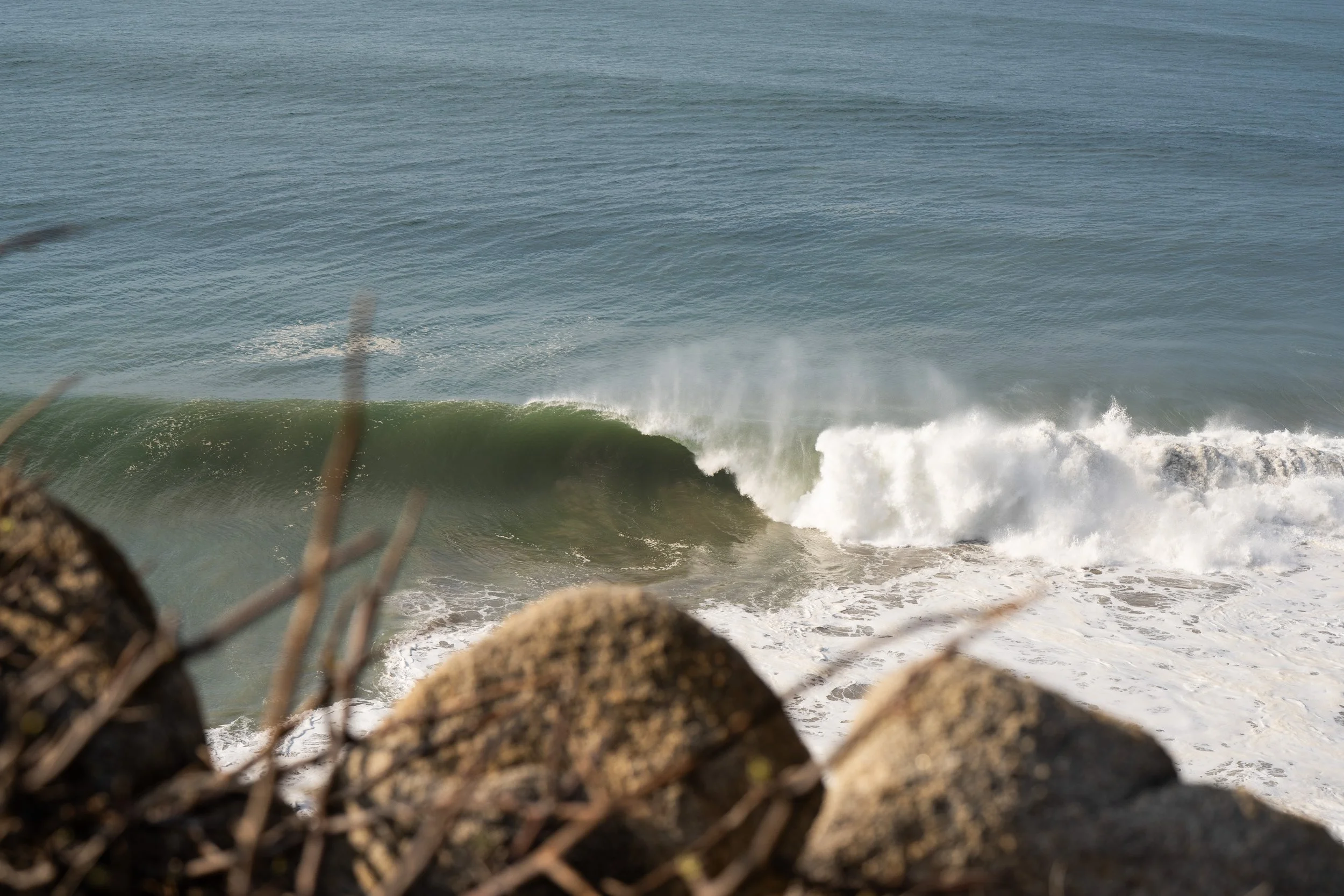 Ocean waves crashing against the rocky shoreline with a chain-link fence in the foreground.