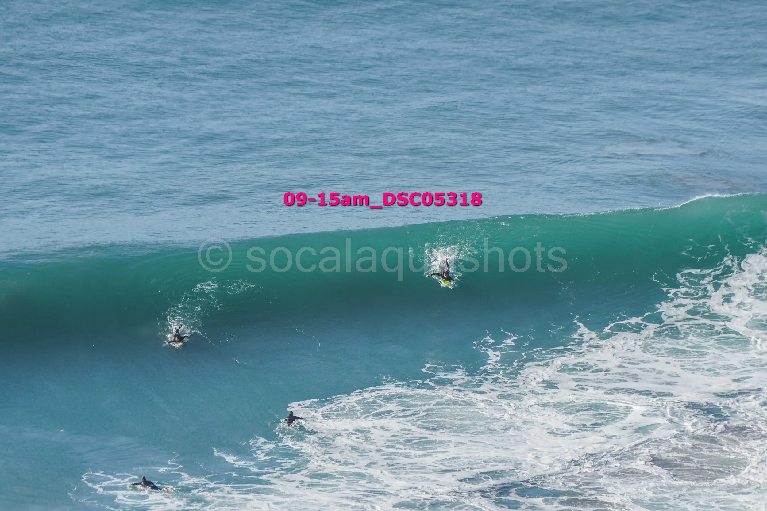 Multiple surfers riding a large ocean wave, with one surfer closer to the wave's crest and others below in the water.
