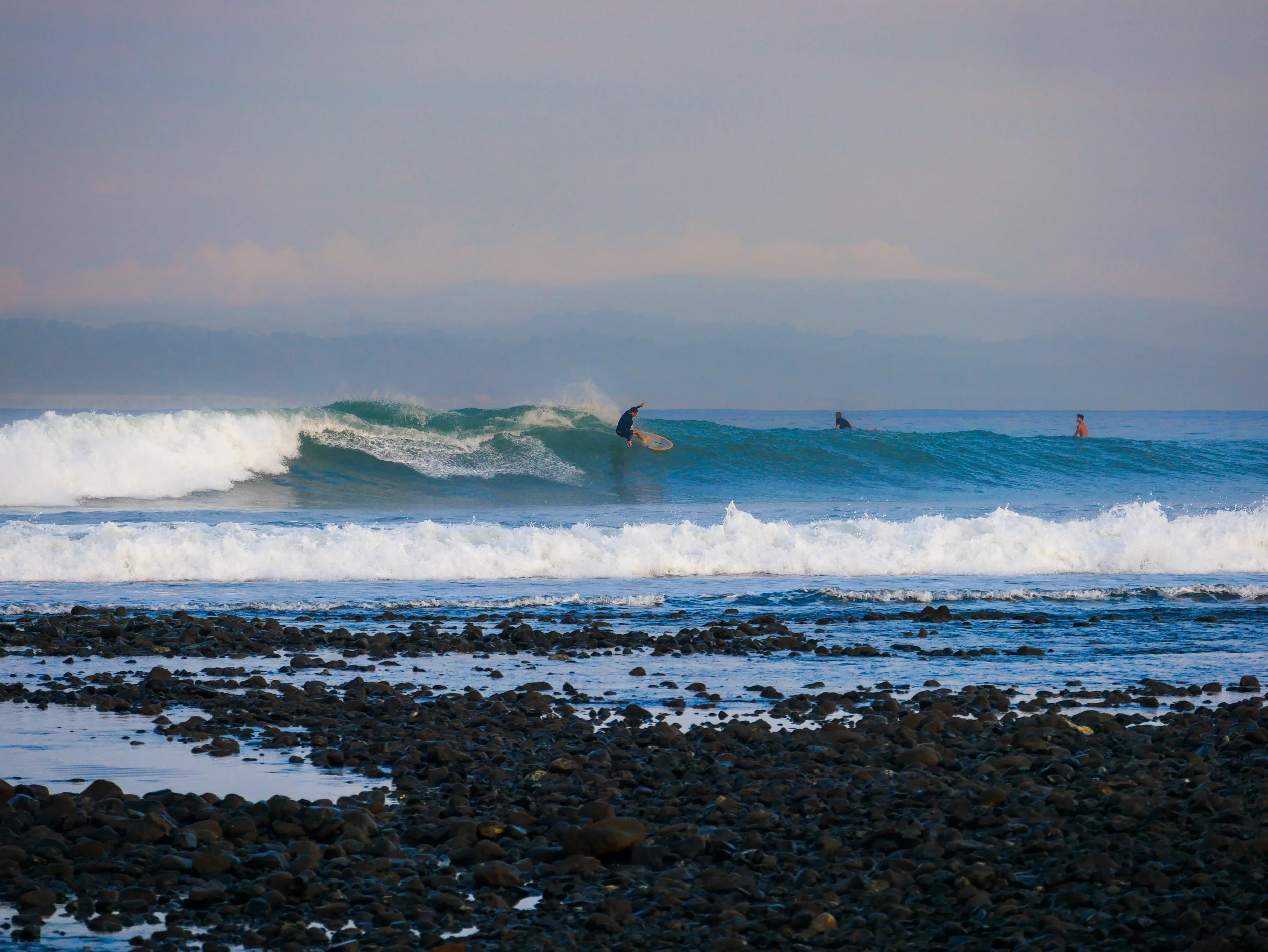 Surfer riding a wave with others waiting in the ocean, rocky shoreline, cloudy sky.