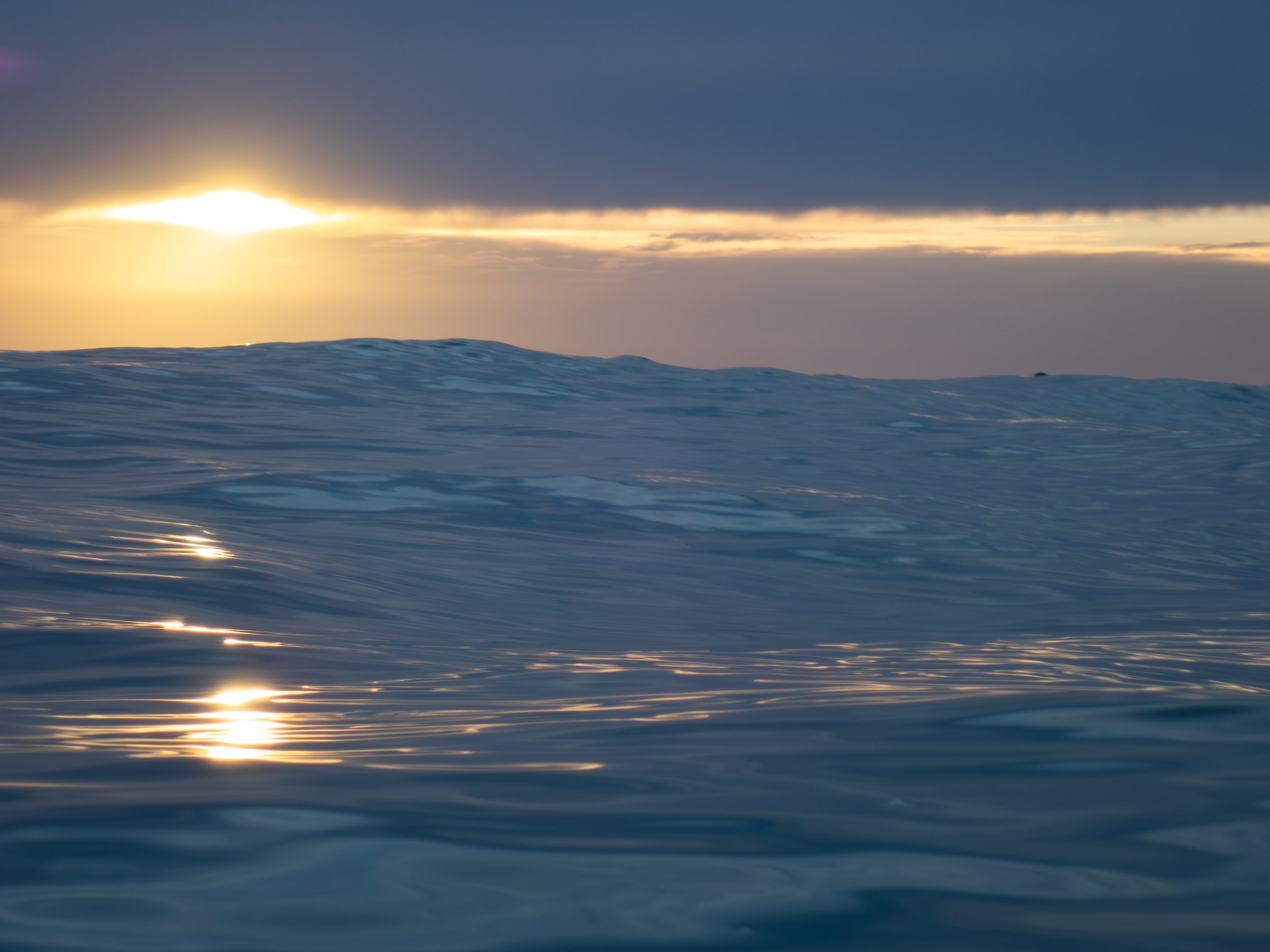 Photo of ocean waves at sunset, with dark clouds and the sun partially visible near the horizon.