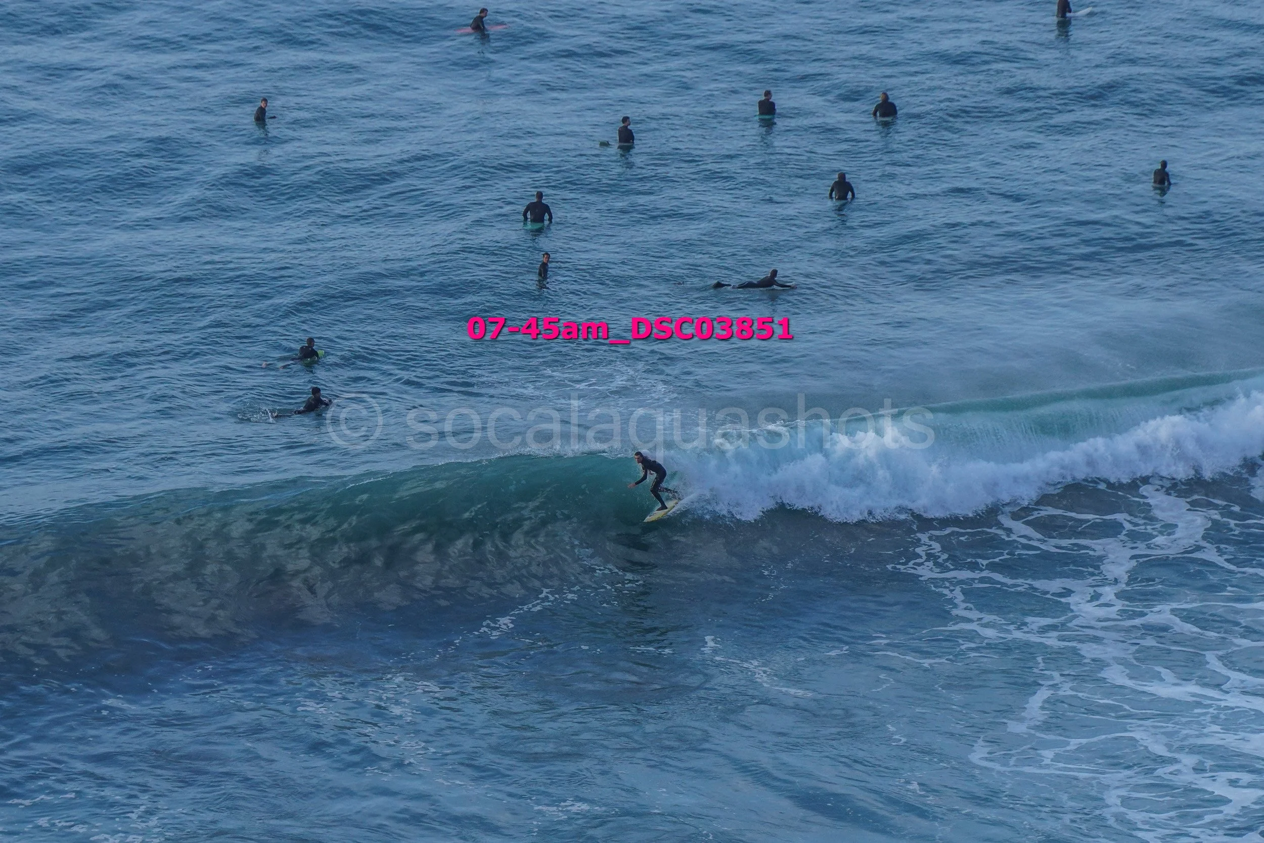 A surfer riding a wave with multiple people swimming and surfing in the water in the background.