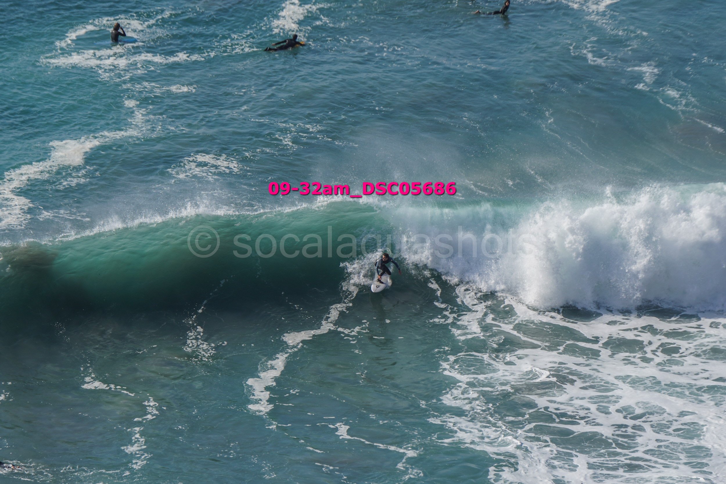 Surfer riding a large wave with several other surfers in the water nearby.