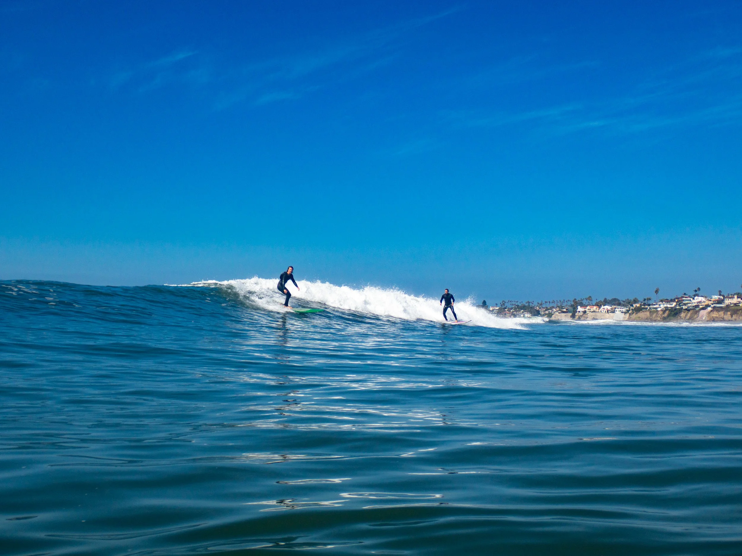 Two surfers riding a wave in the ocean with a clear blue sky and a coastline with buildings and palm trees in the background.