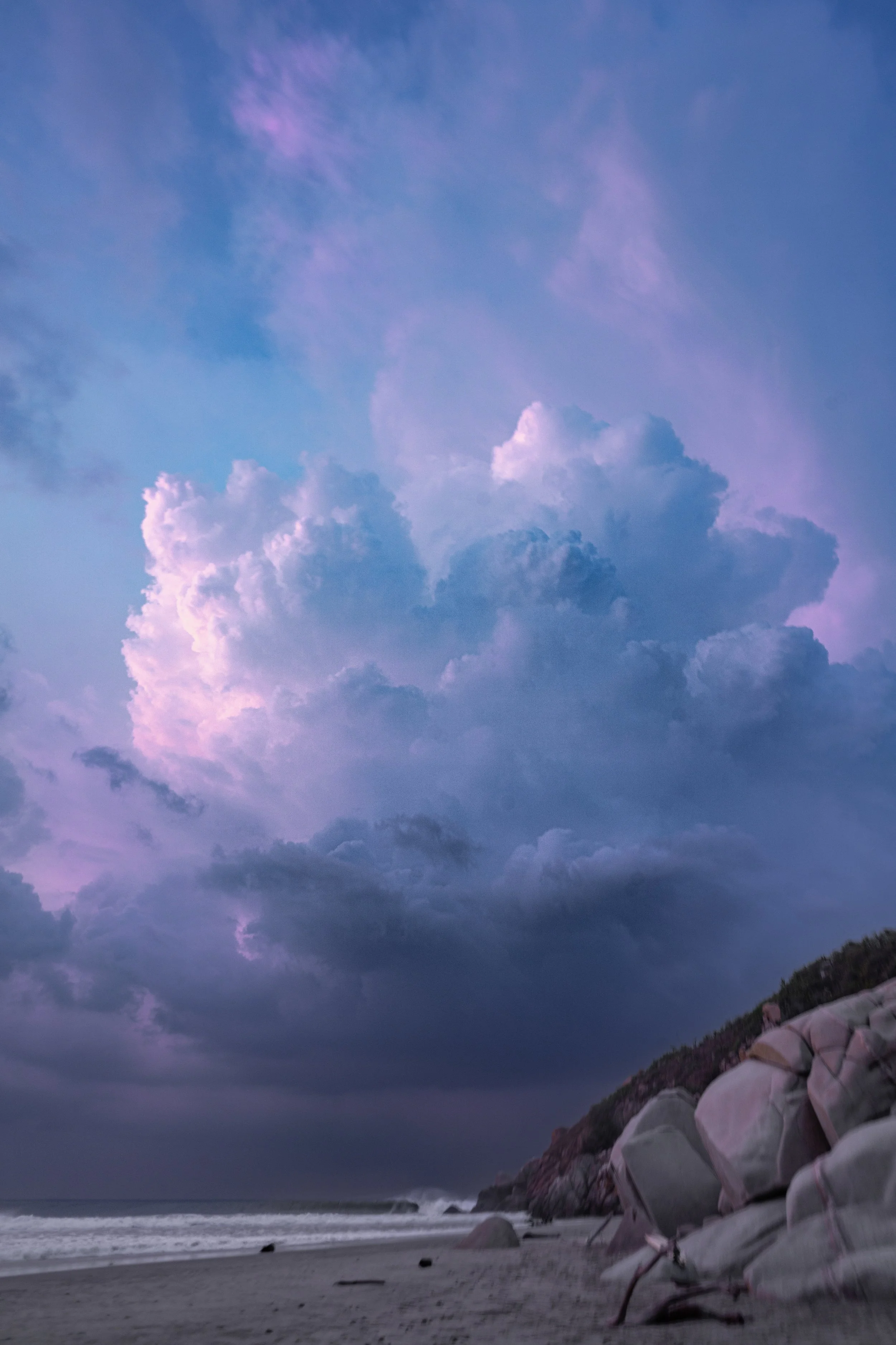 A beach scene at dusk with rocky shoreline, ocean waves, and cloudy sky with purple and pink hues.